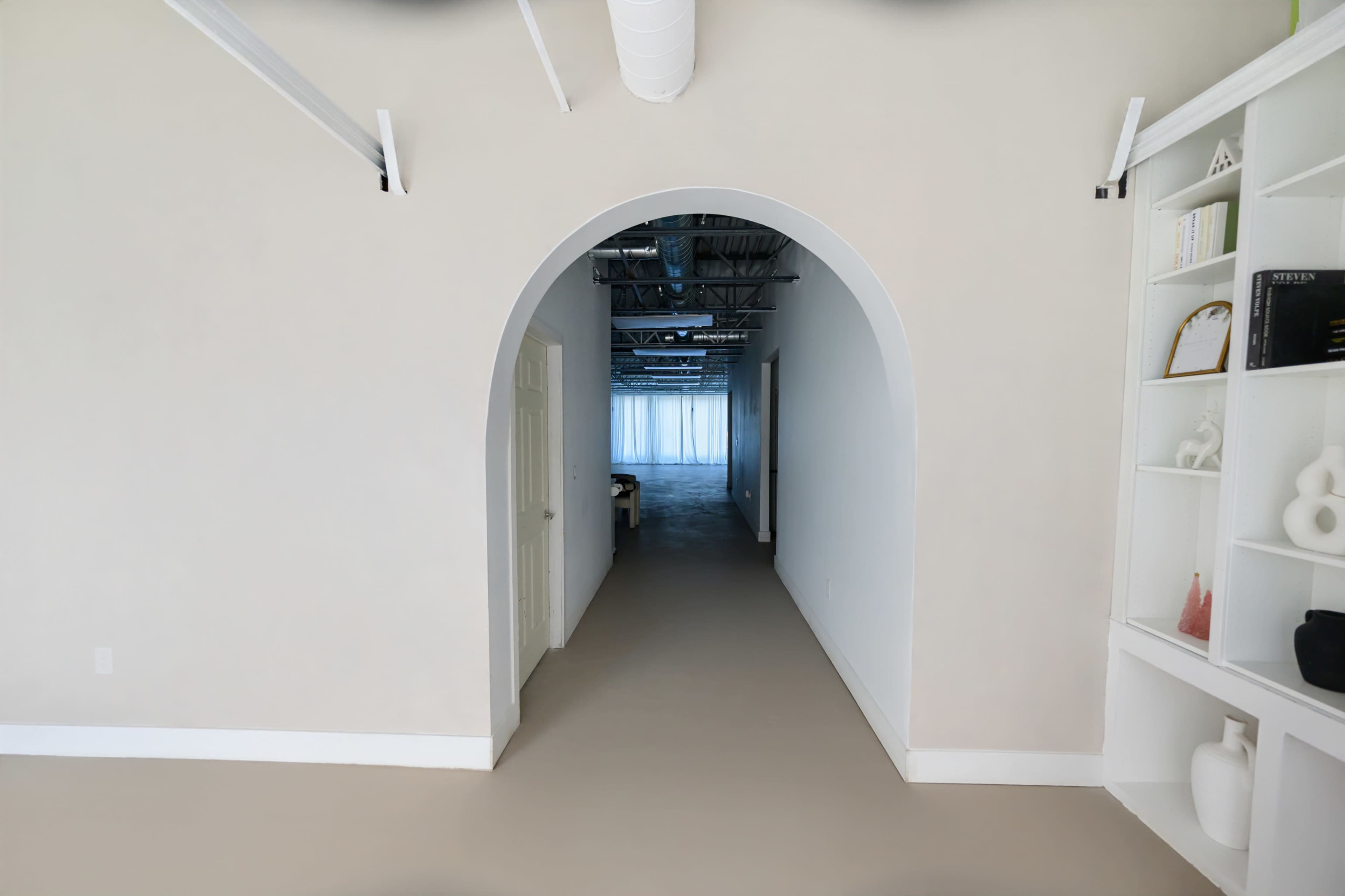 The image shows a white-walled corridor leading through an archway, with a shelf on the right displaying various decorative objects.