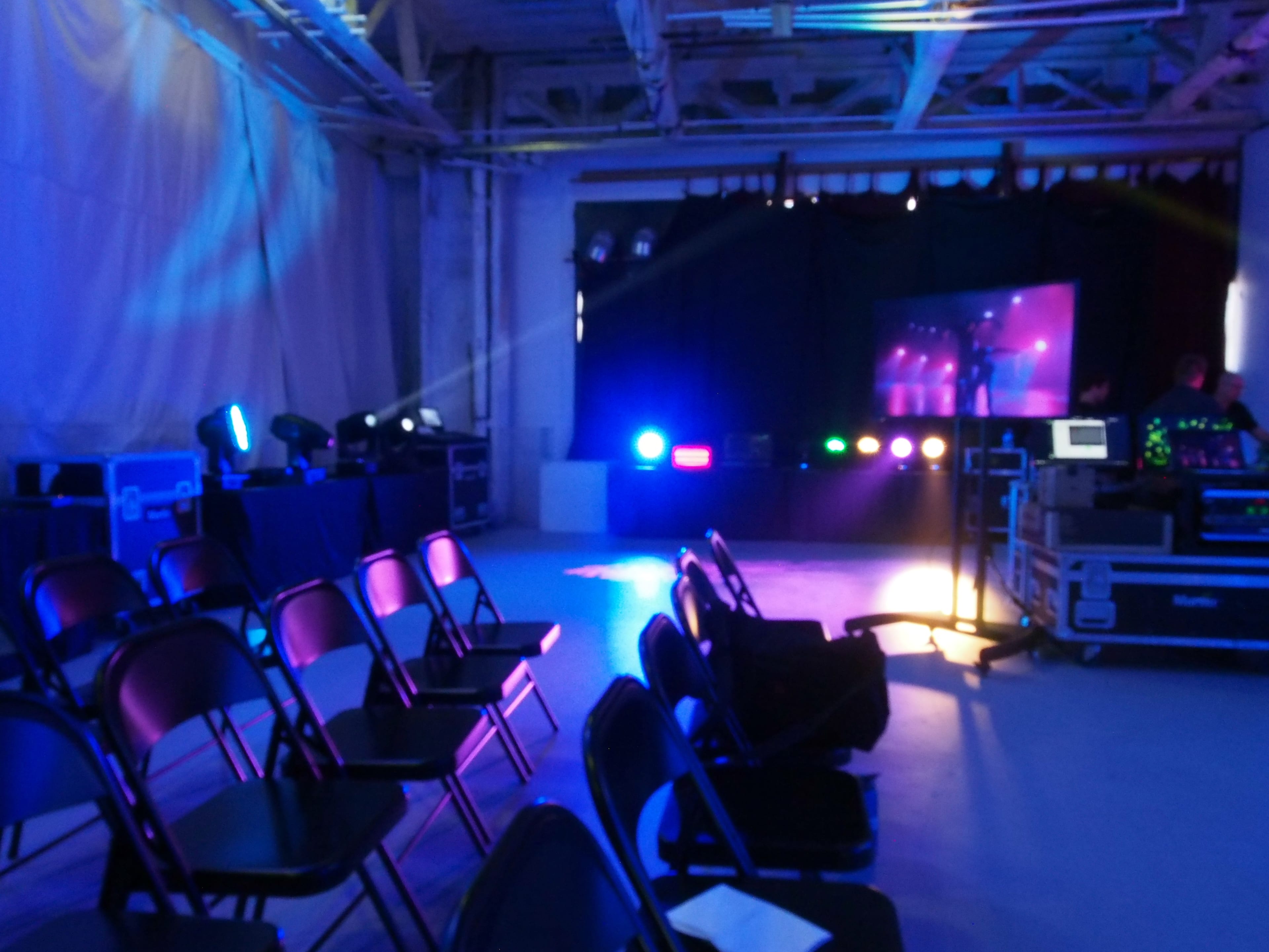 An indoor event space with a row of black chairs facing a stage illuminated by colorful lights and a screen.
