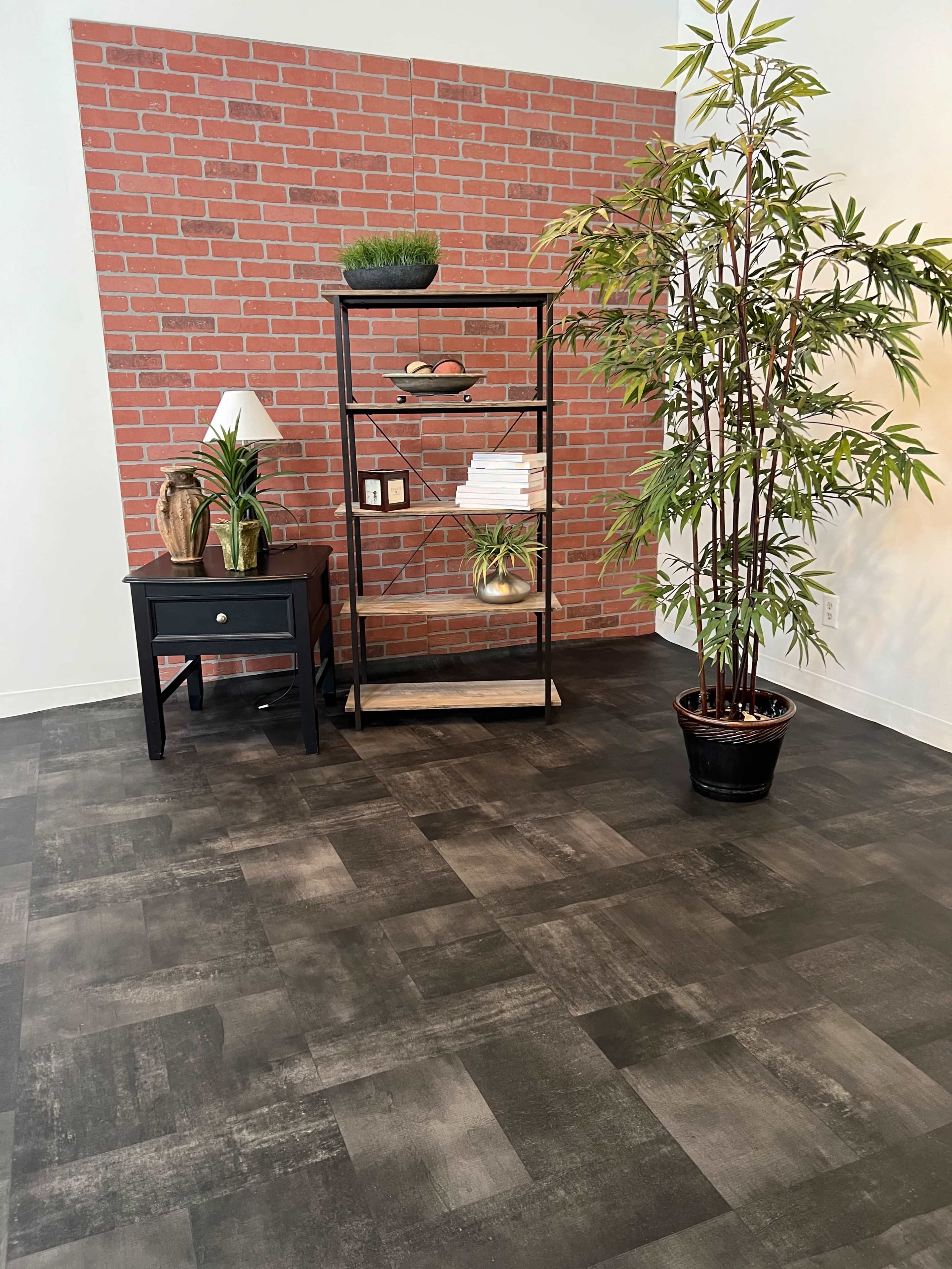 A corner of a room featuring a red brick accent wall, a black shelf with decorative items, a small black nightstand, and a potted plant in the foreground.