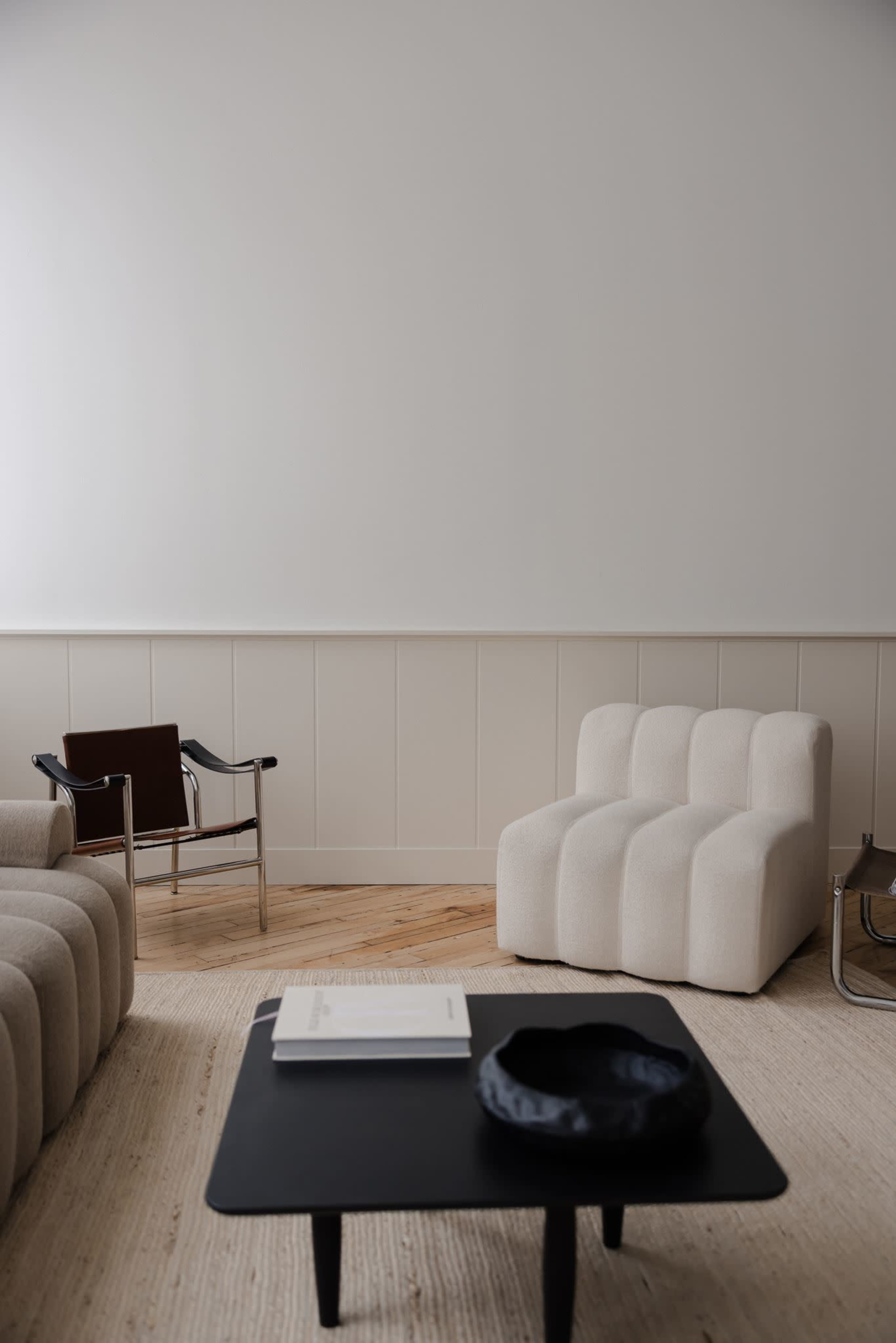 The image shows a minimalistic living room with a plush white chair, a beige sofa, a black coffee table, and a light-colored rug on a wooden floor.