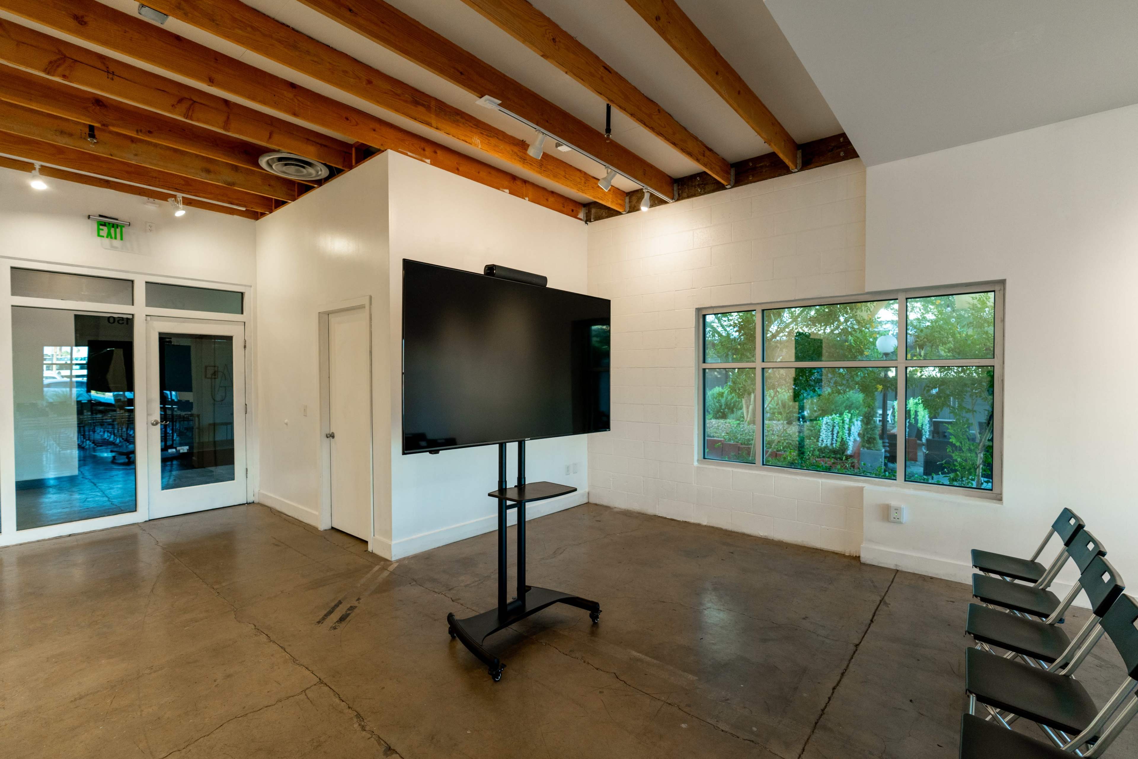 A spacious room features a large mounted screen on a stand, with several chairs arranged nearby and a window providing natural light.