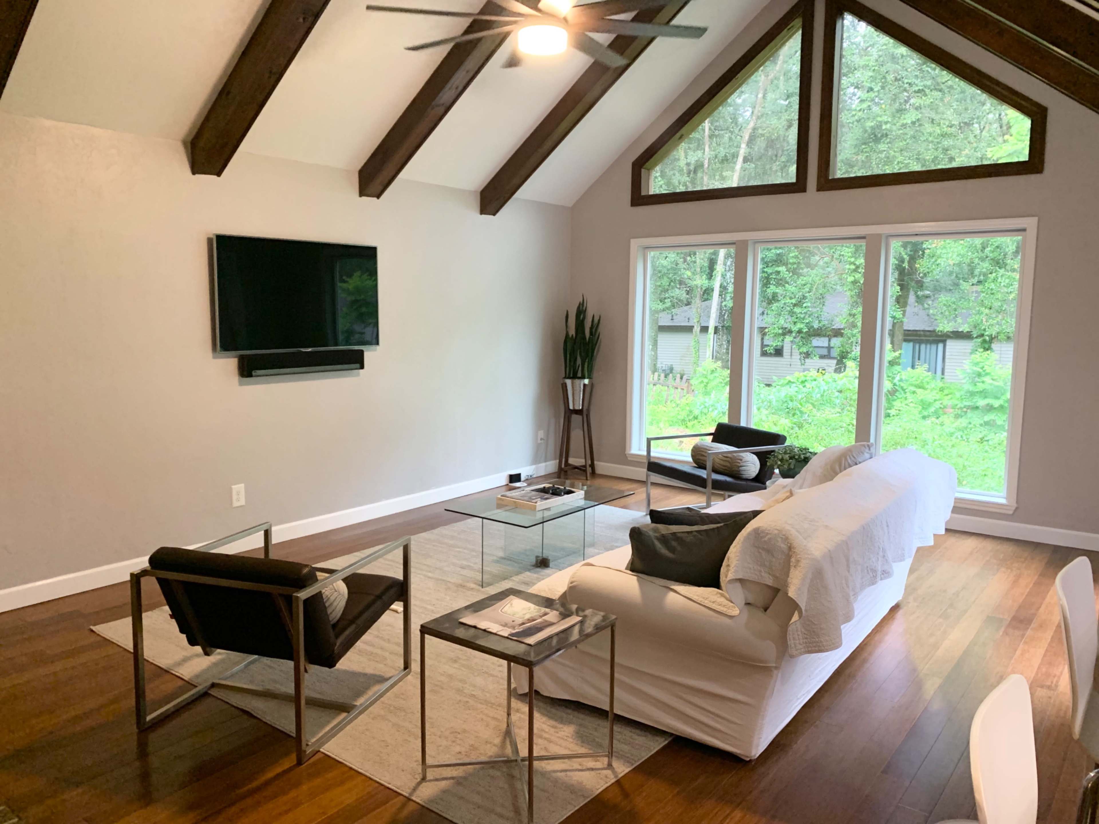 The living room features a white sofa, two black chairs, a glass coffee table, and large windows overlooking a green landscape.