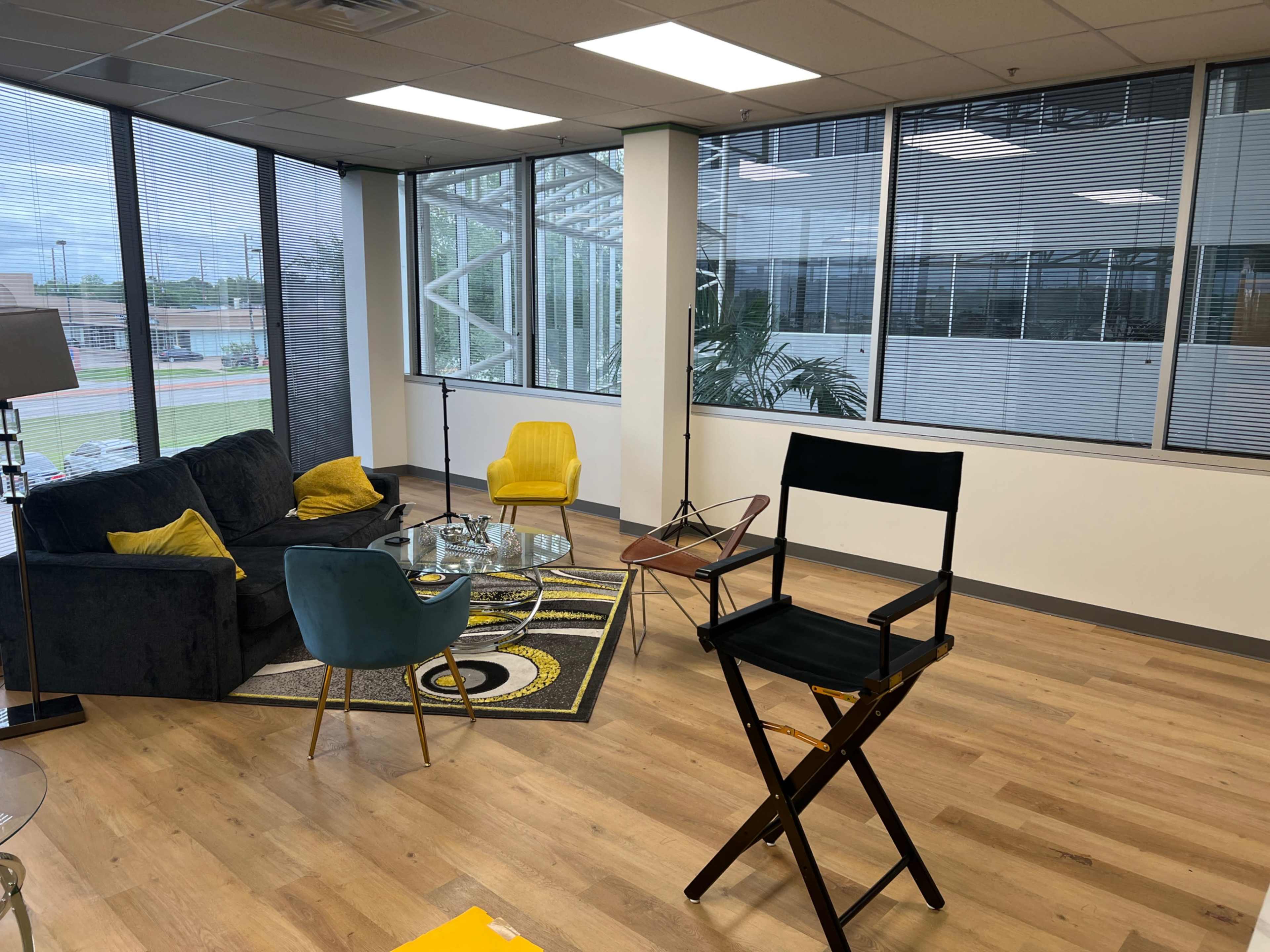 A well-lit office space featuring a dark couch, colorful armchairs, a glass coffee table, and a director's chair, with large windows allowing natural light to enter.