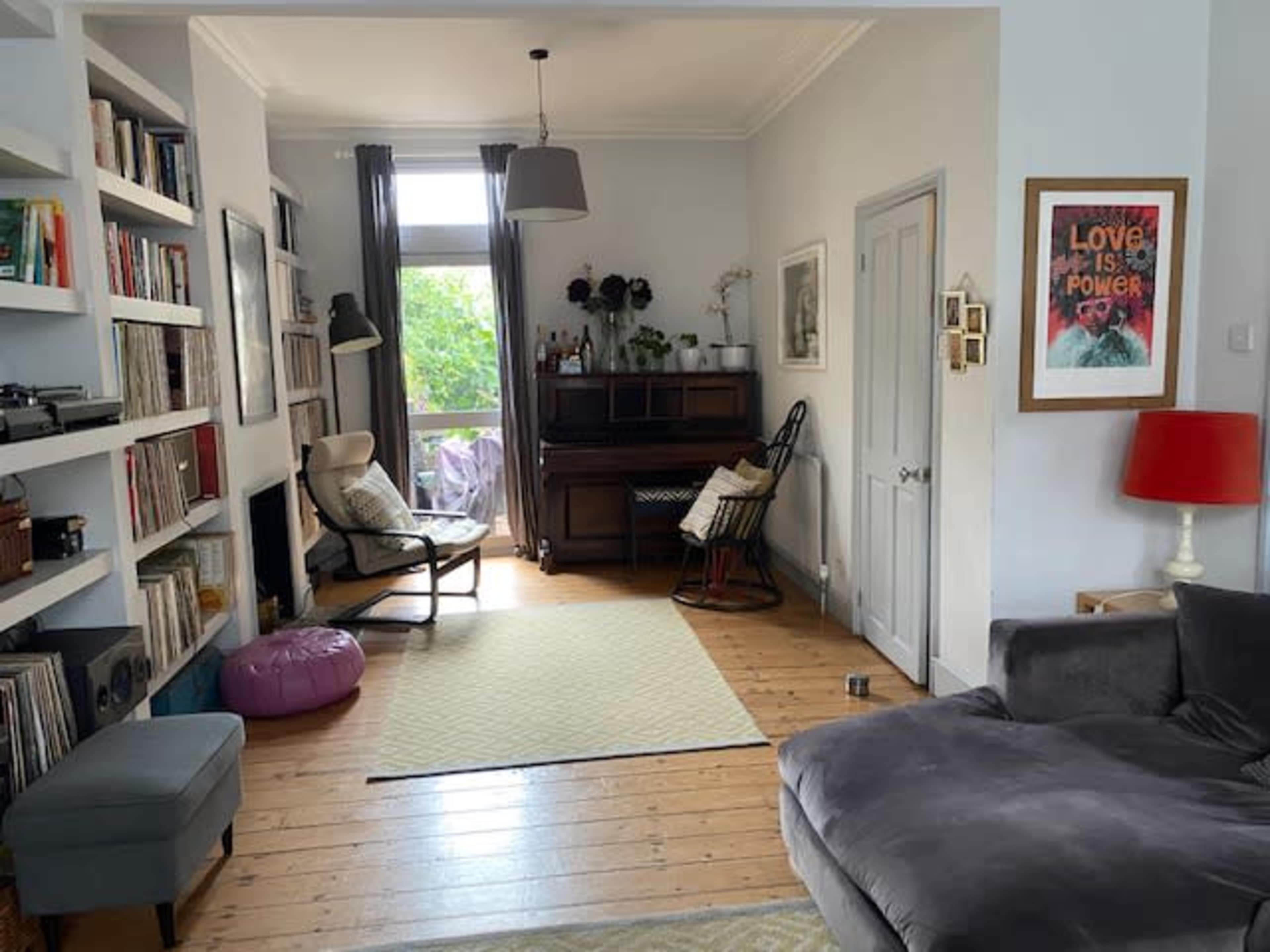 The room features a cozy seating area with a bookshelf, a piano, and large windows allowing natural light.