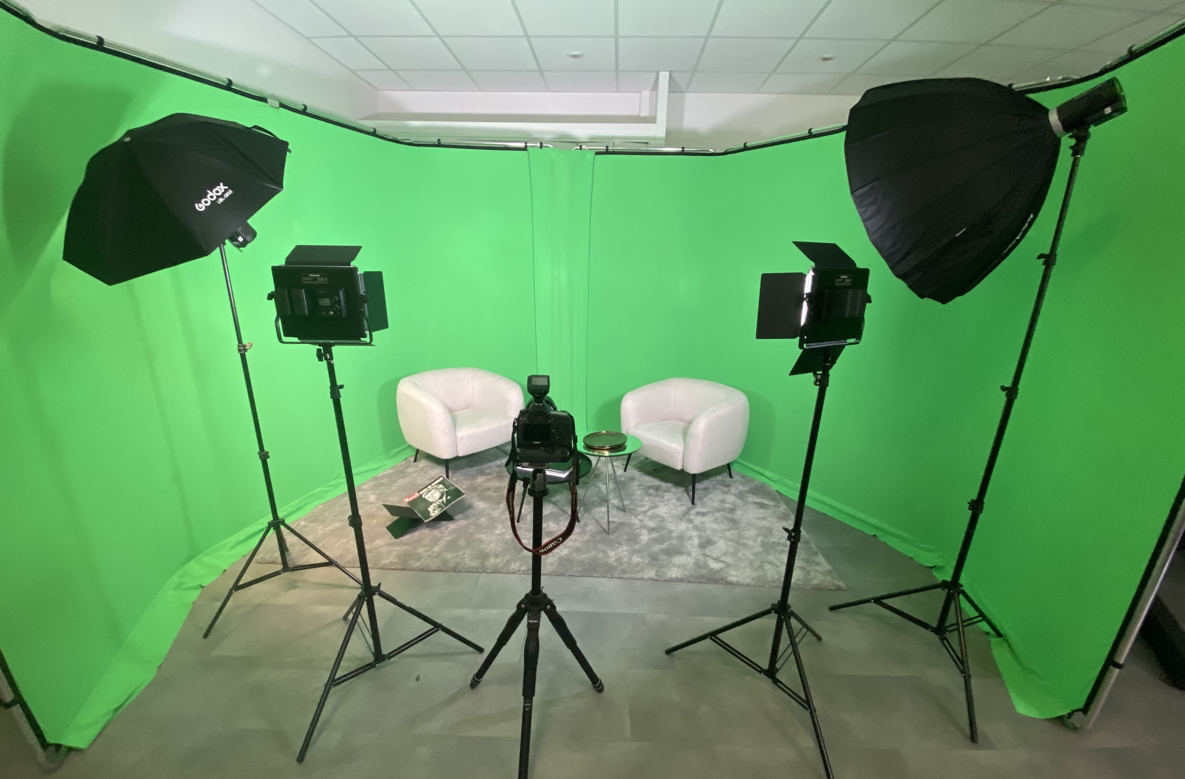 The image shows a green screen setup featuring two white chairs, a small table, and studio lighting equipment arranged around a camera on a tripod.