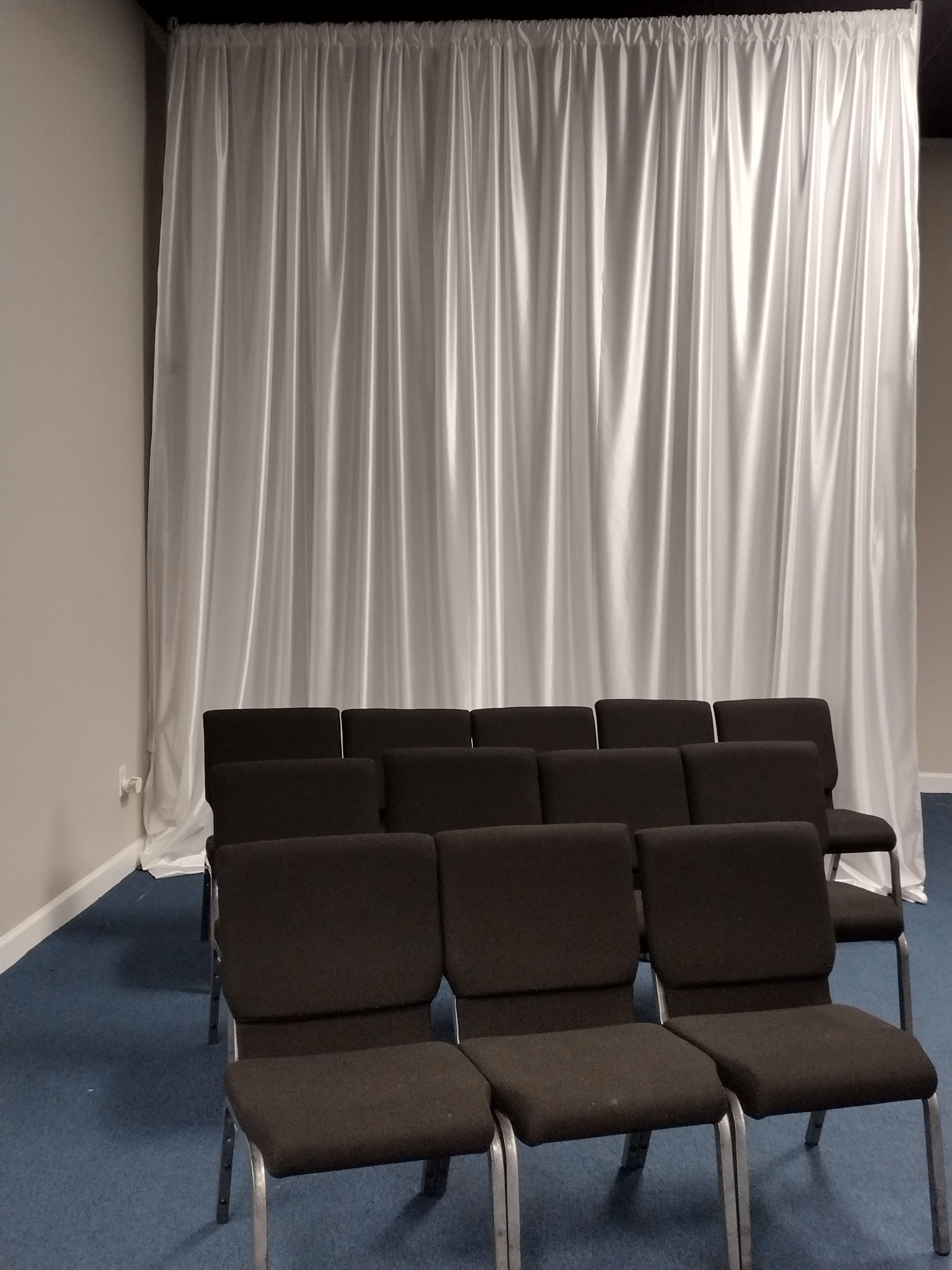A row of black chairs is set against a backdrop of white drapery in a room with blue carpeting.