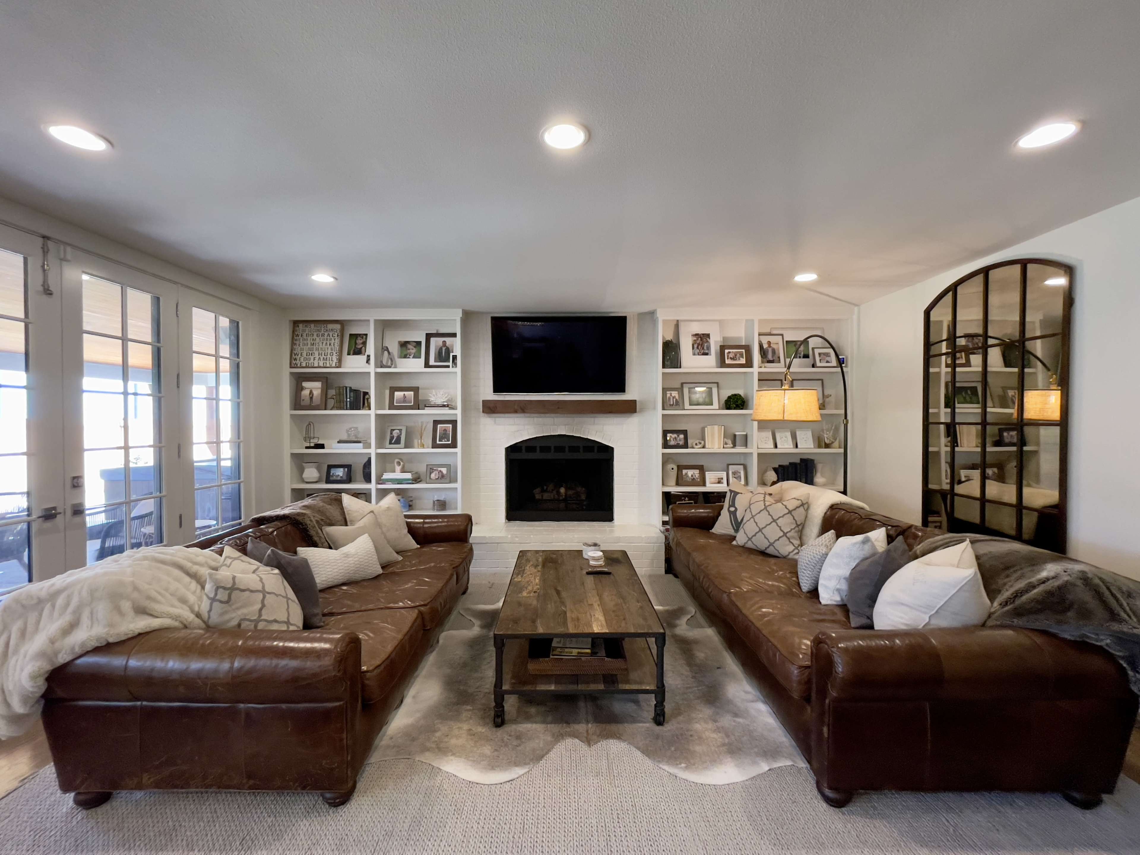 A living room features two leather sofas facing a coffee table, with a fireplace and built-in shelves displaying framed photographs and decorative items.