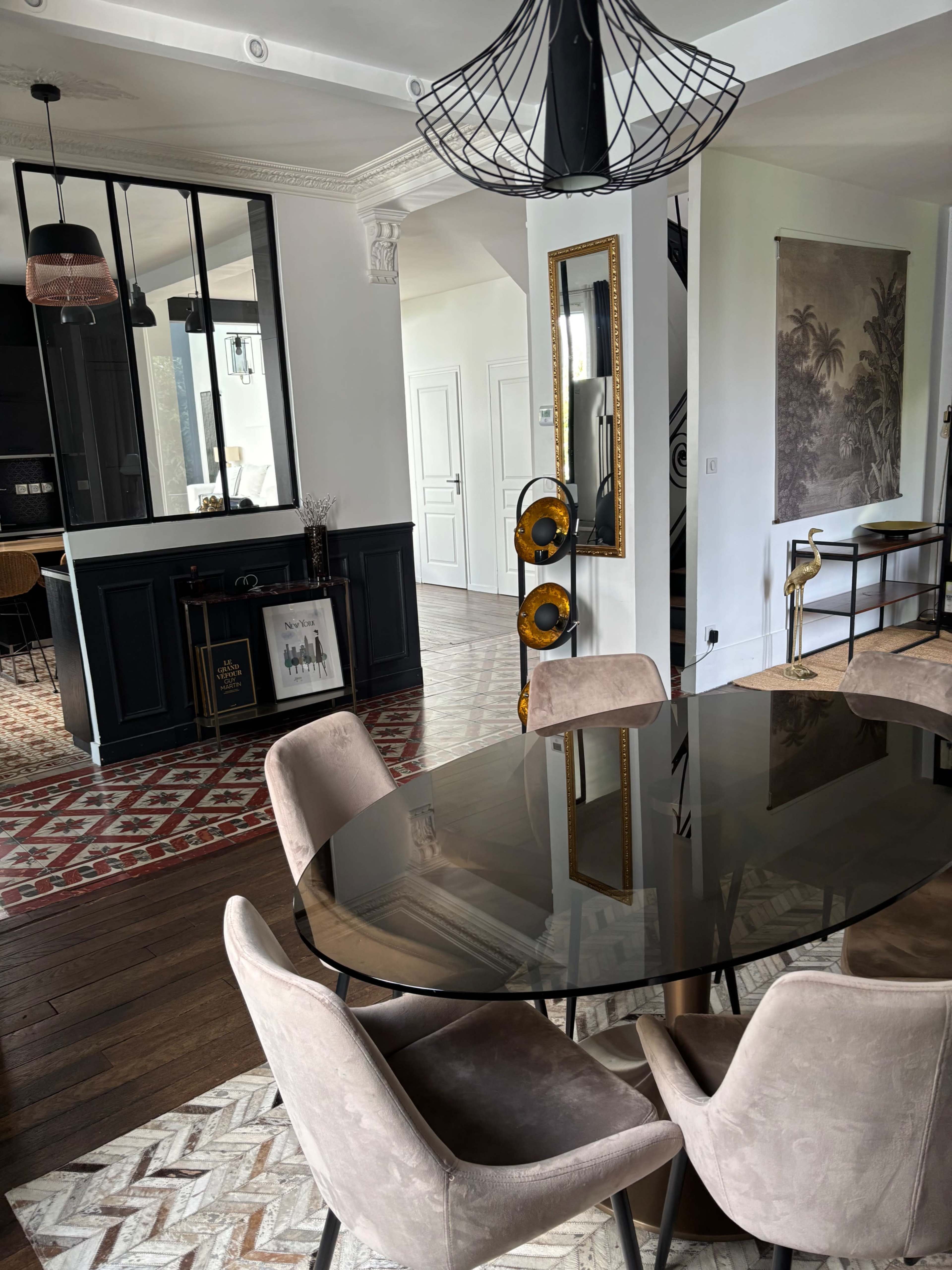 A modern dining area features a round glass table surrounded by upholstered chairs, with a view of an open kitchen and decorative elements in the background.