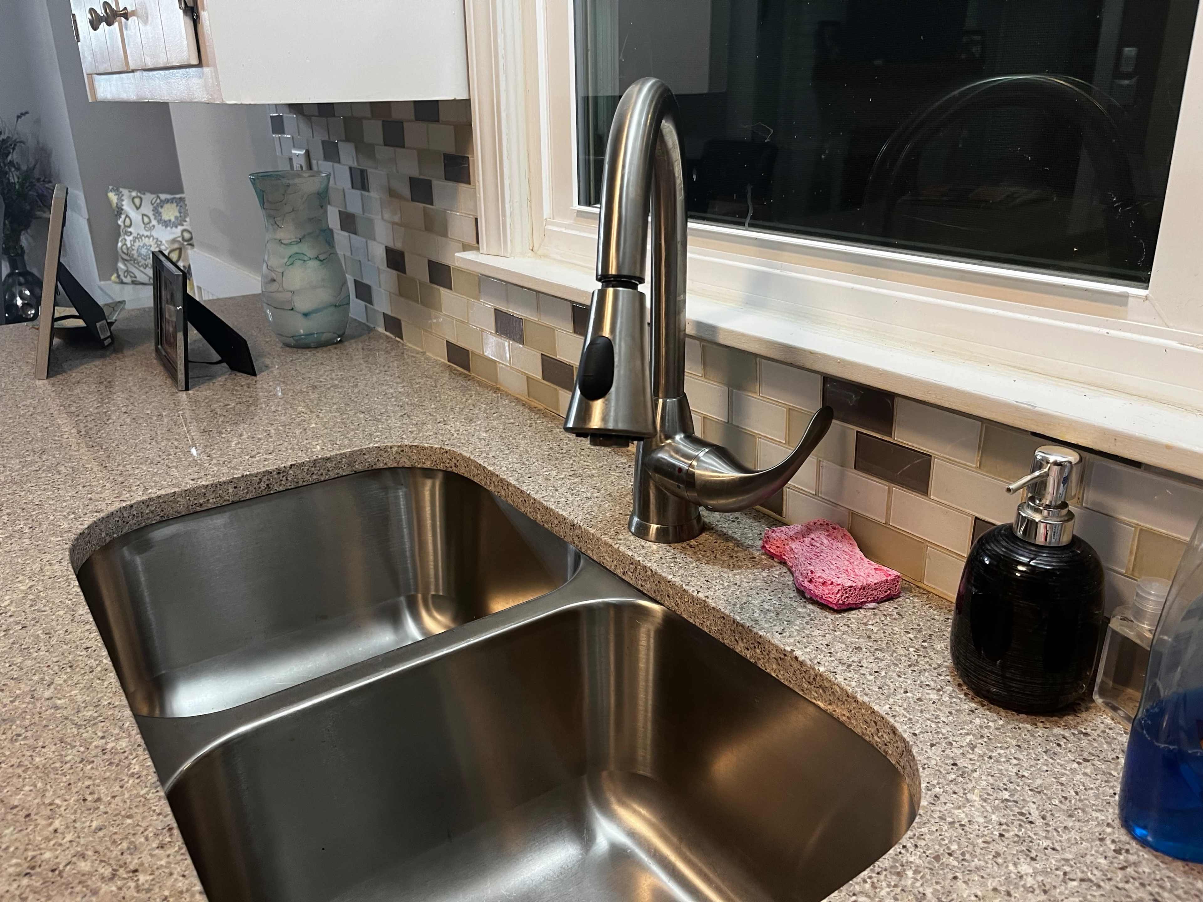 A kitchen sink with a stainless steel faucet, a soap dispenser, and a pink sponge on the counter beside it, all set against a tiled backsplash.