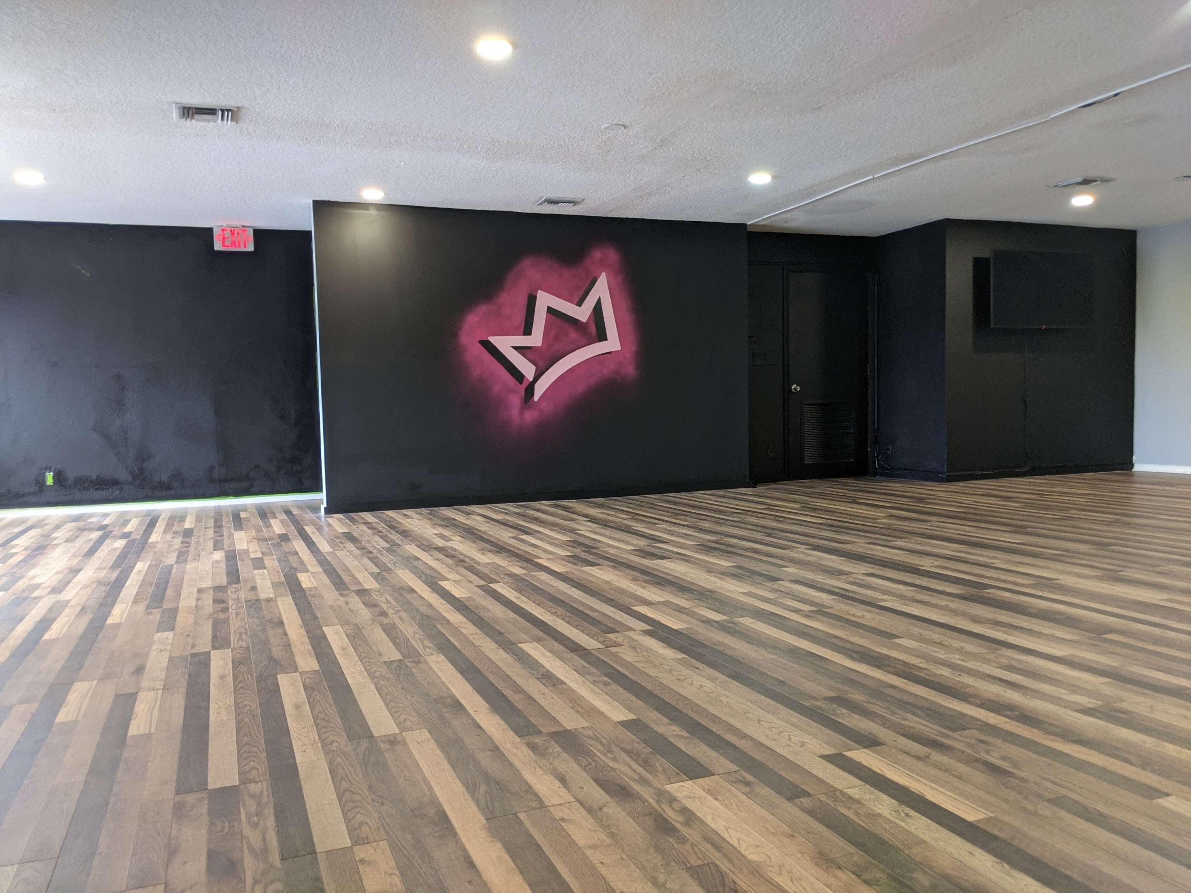 An empty studio space with wooden flooring and a black wall featuring a pink crown graphic.