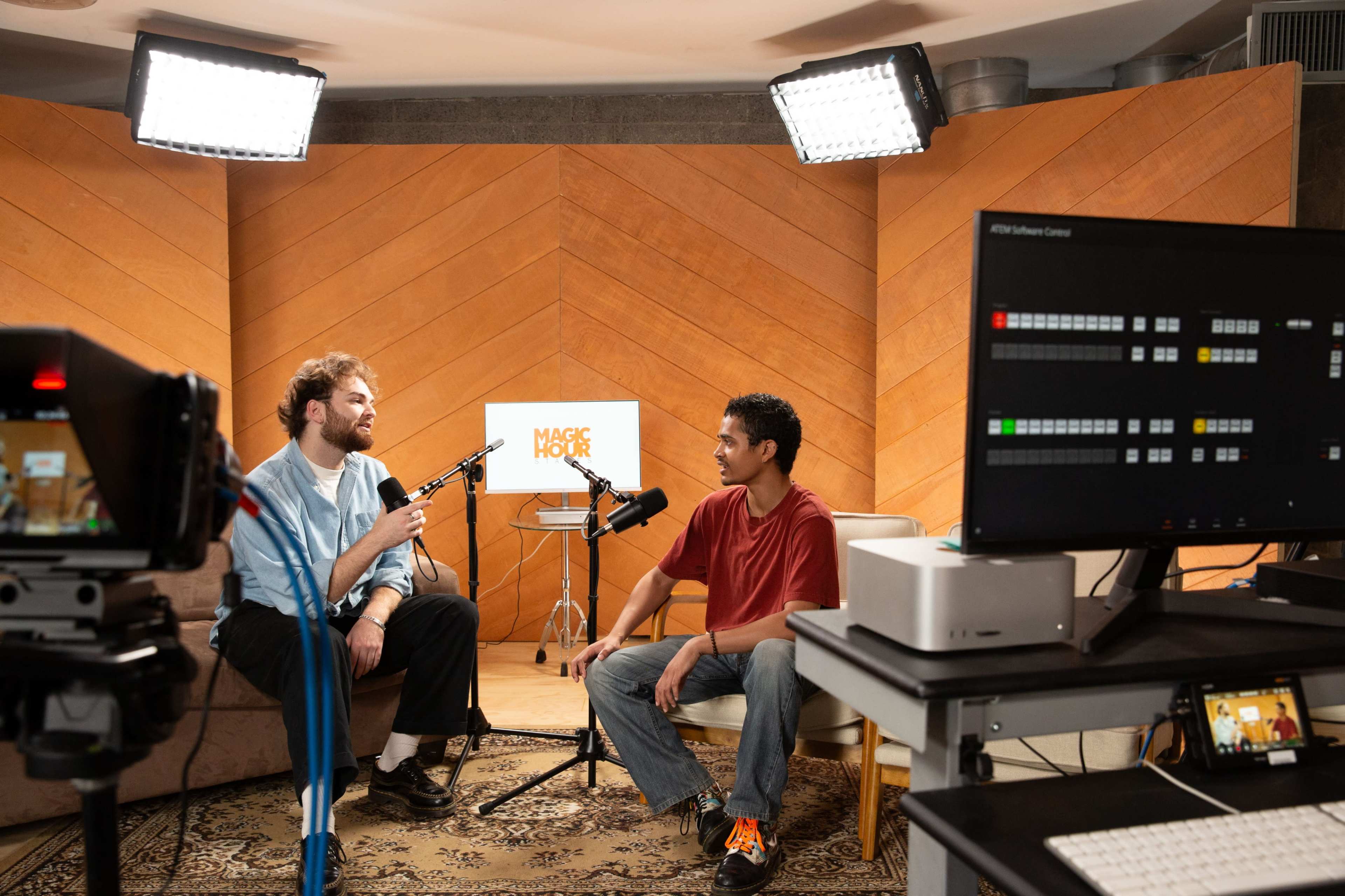 Two men sit in a well-lit studio, speaking into microphones while cameras and monitors capture the conversation.