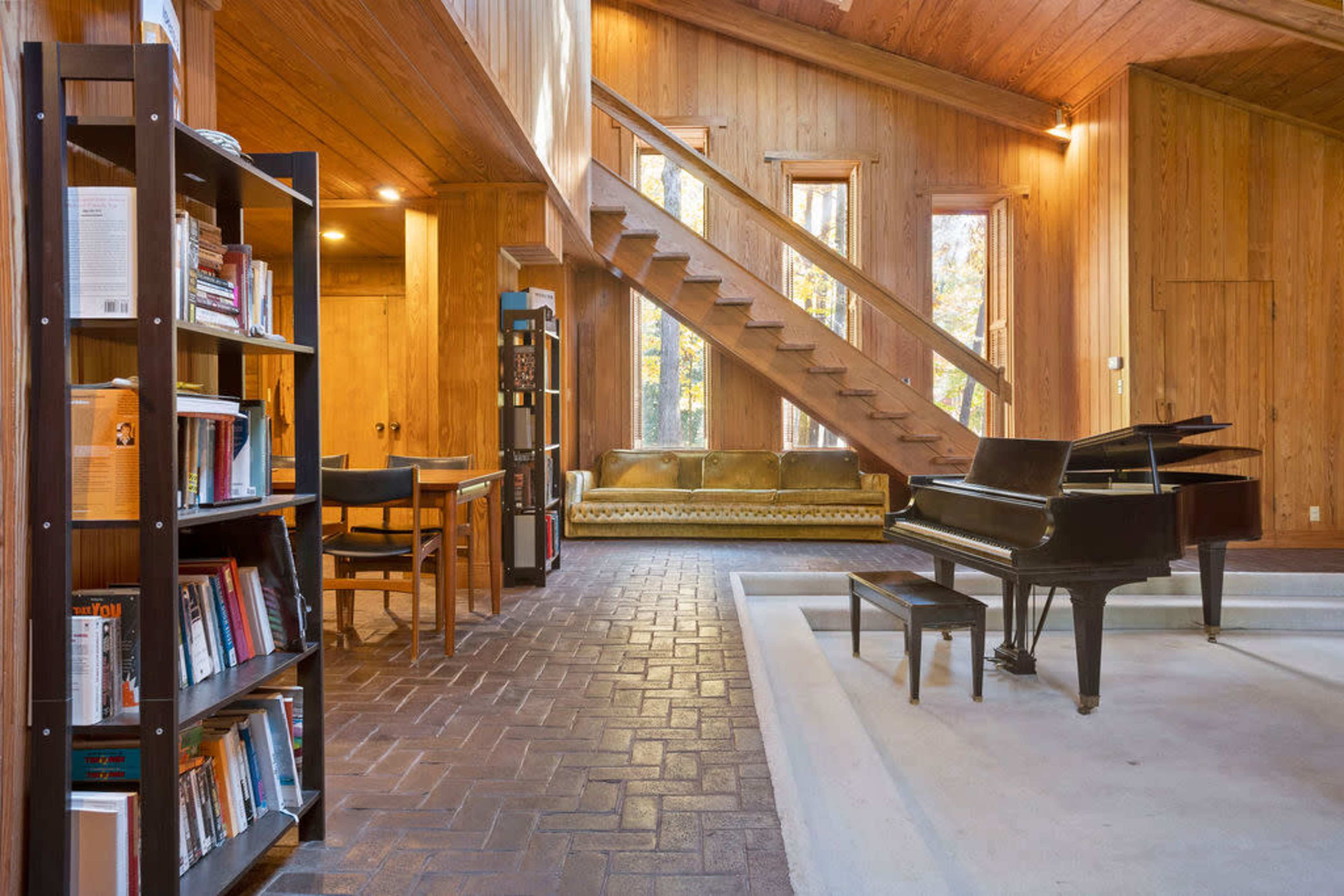 A spacious wooden interior with a staircase, a grand piano, a bookshelf filled with books, a seating area, and large windows allowing natural light.