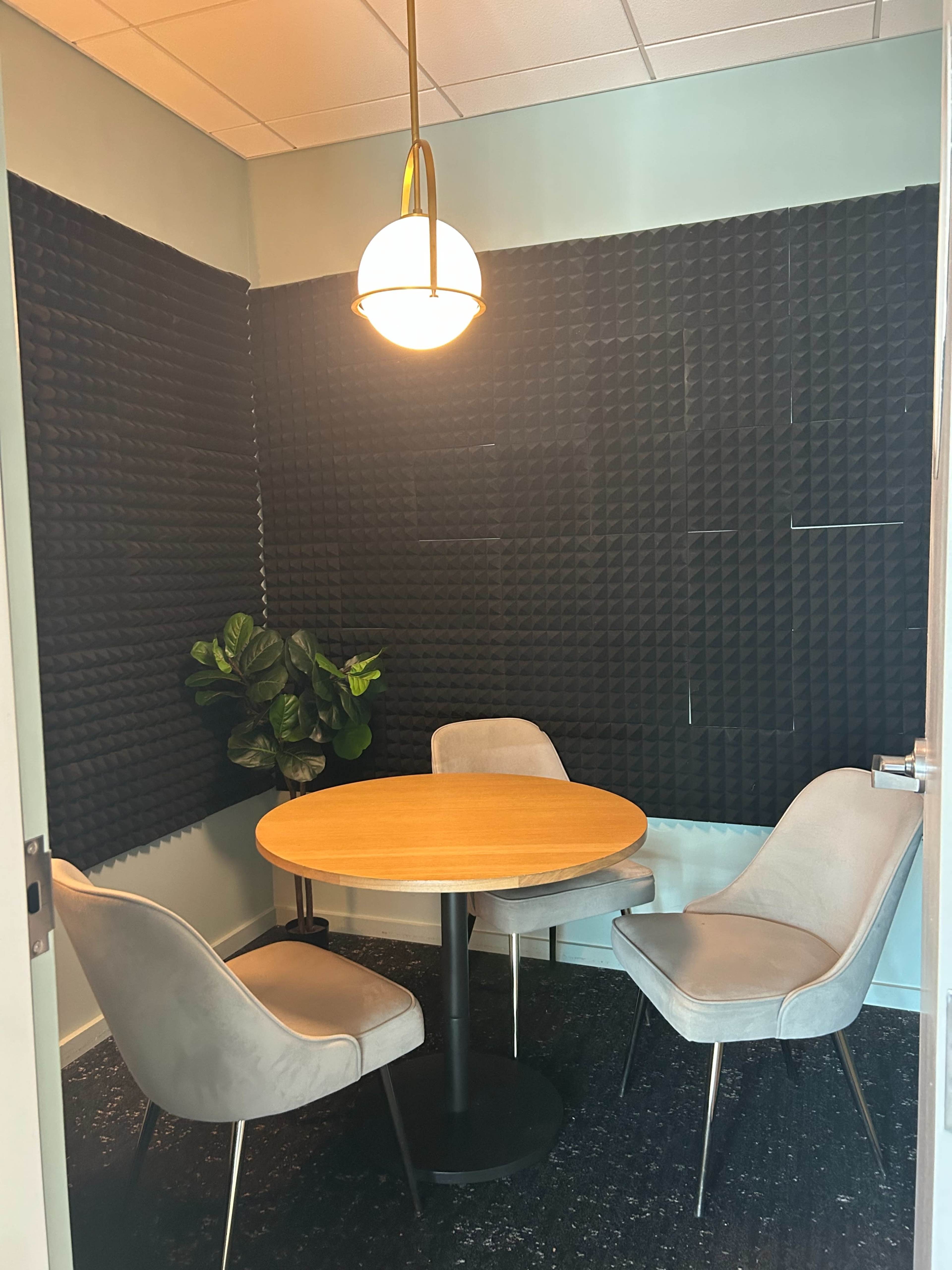 The image shows a small meeting room with a round wooden table surrounded by three gray chairs, and textured black soundproofing panels on the walls.
