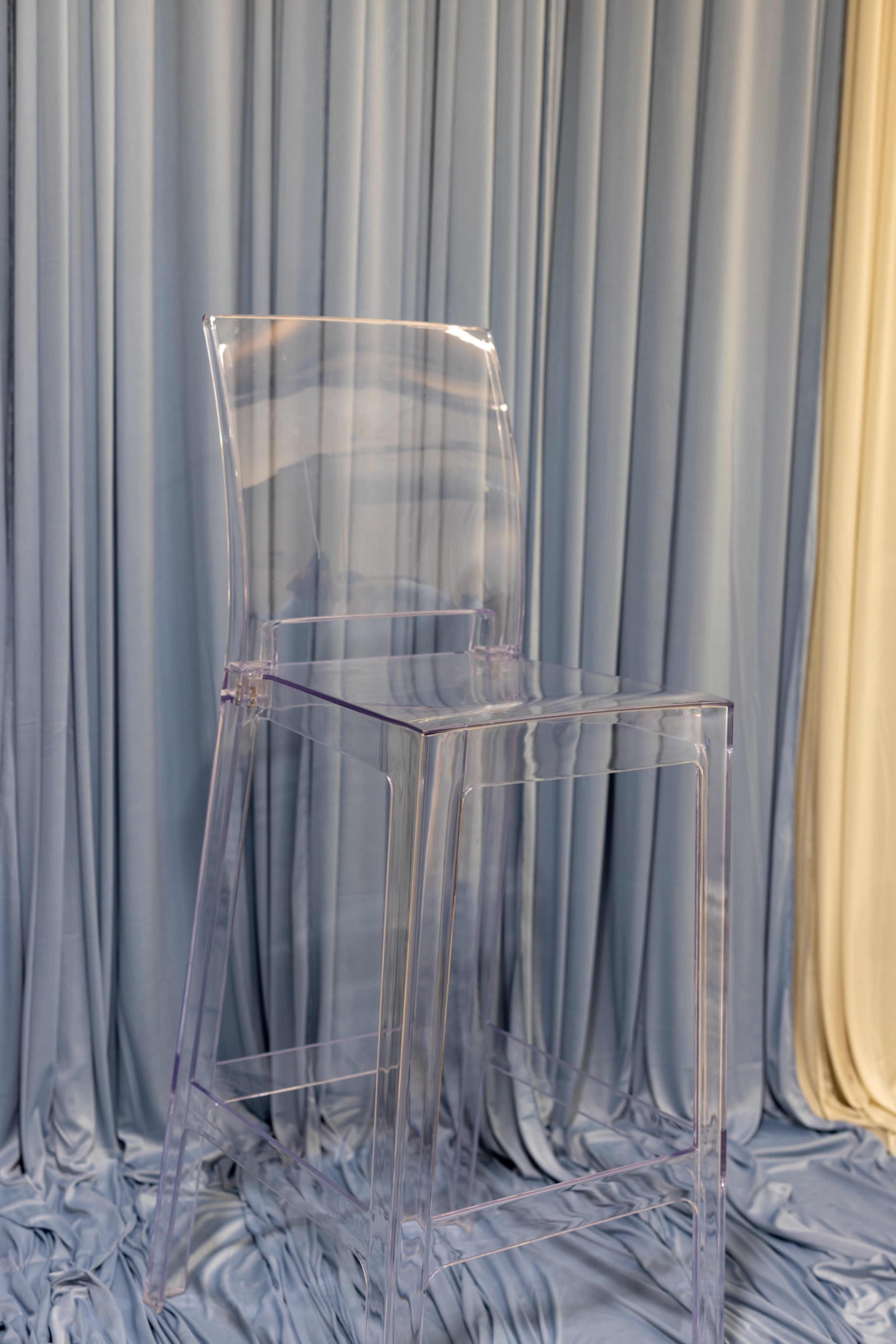 A clear acrylic bar stool is positioned in front of draped fabric in shades of blue and cream.