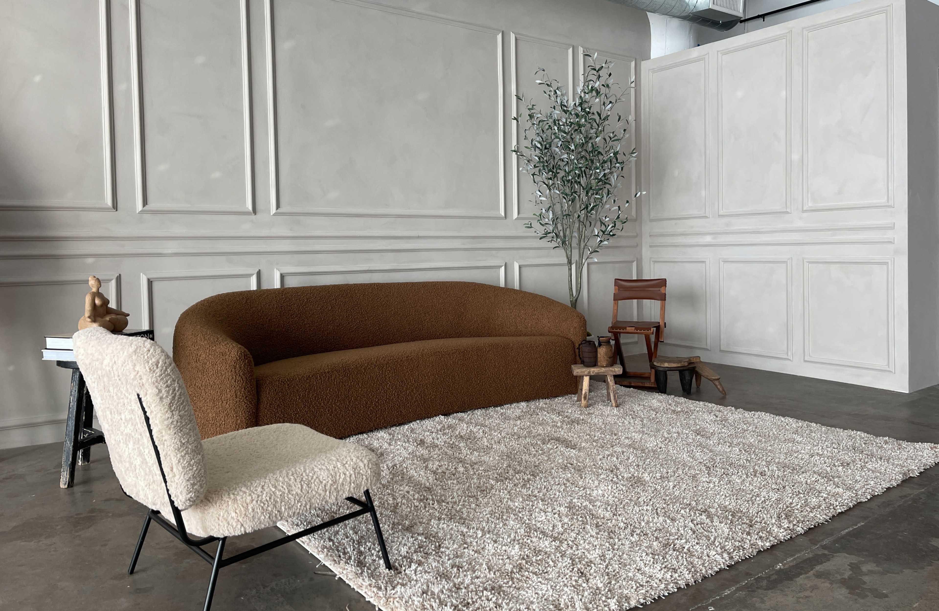 A modern living space featuring a brown curved sofa, a beige armchair, a cream-colored rug, and a decorative tree in a minimalist setting.
