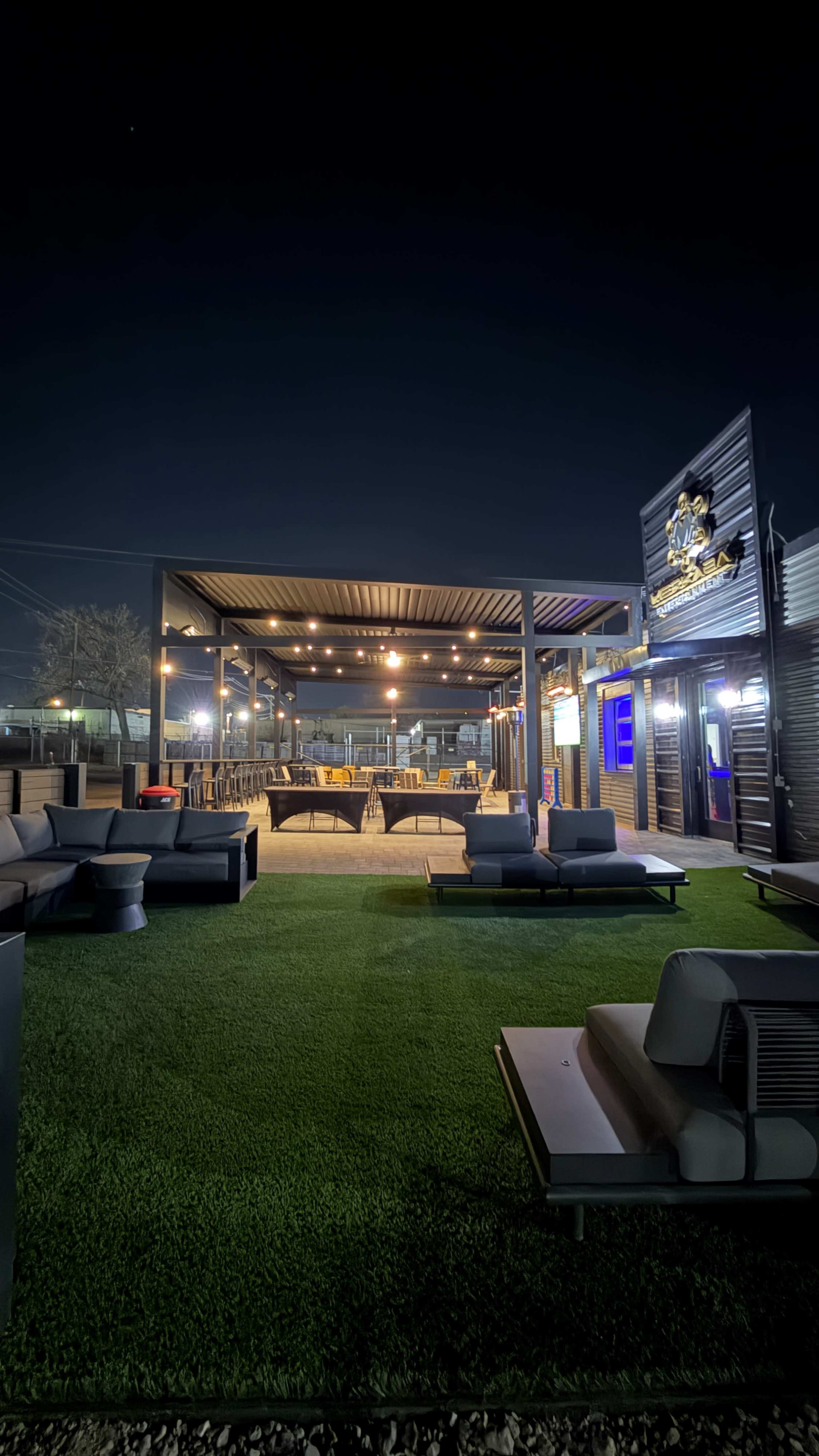 The image shows a modern outdoor seating area with sleek furniture, green grass, and string lights, set against a night sky.
