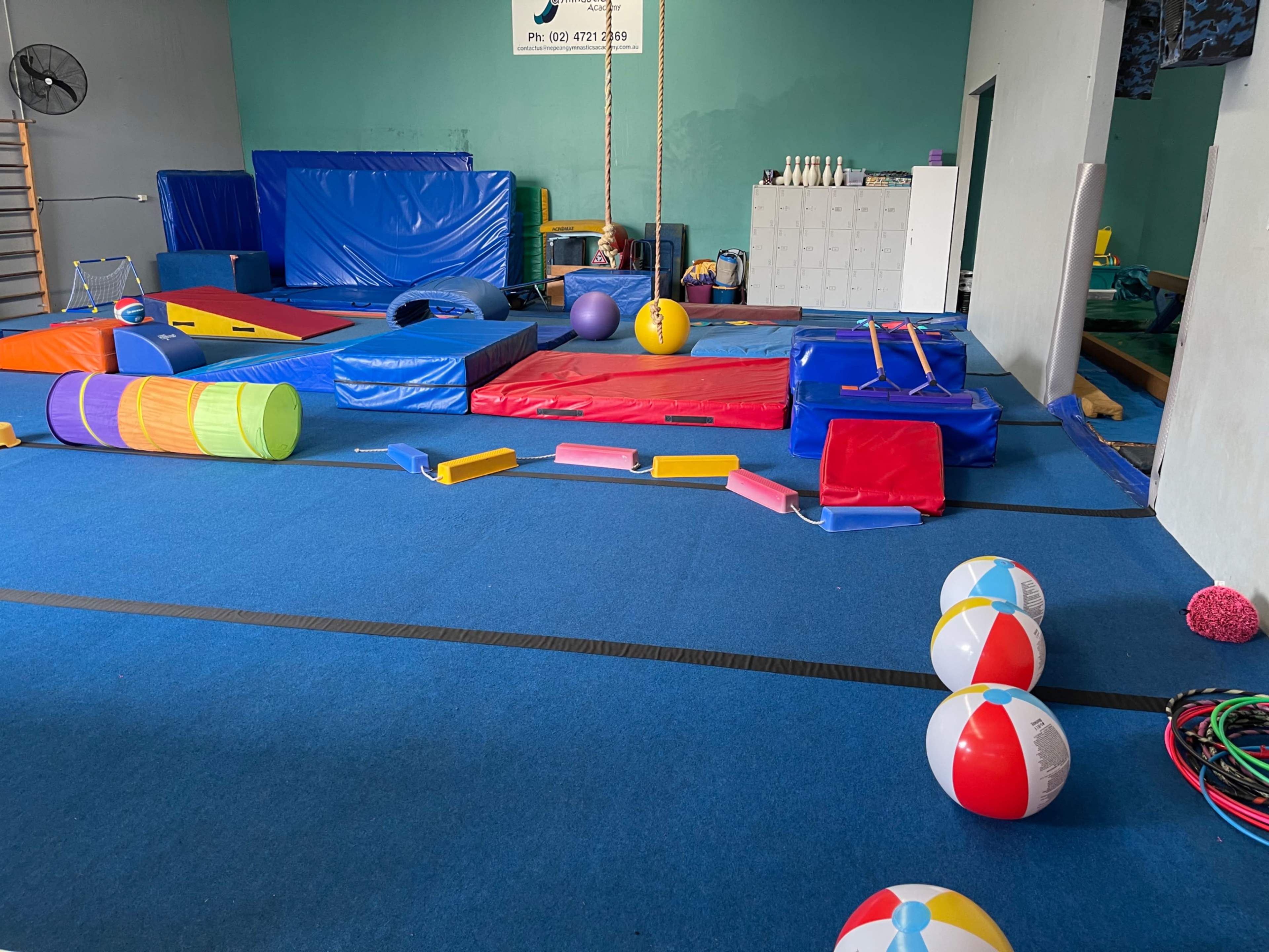 A brightly colored gymnastics or play area filled with various mats, equipment, and exercise balls arranged on a blue rubber floor.