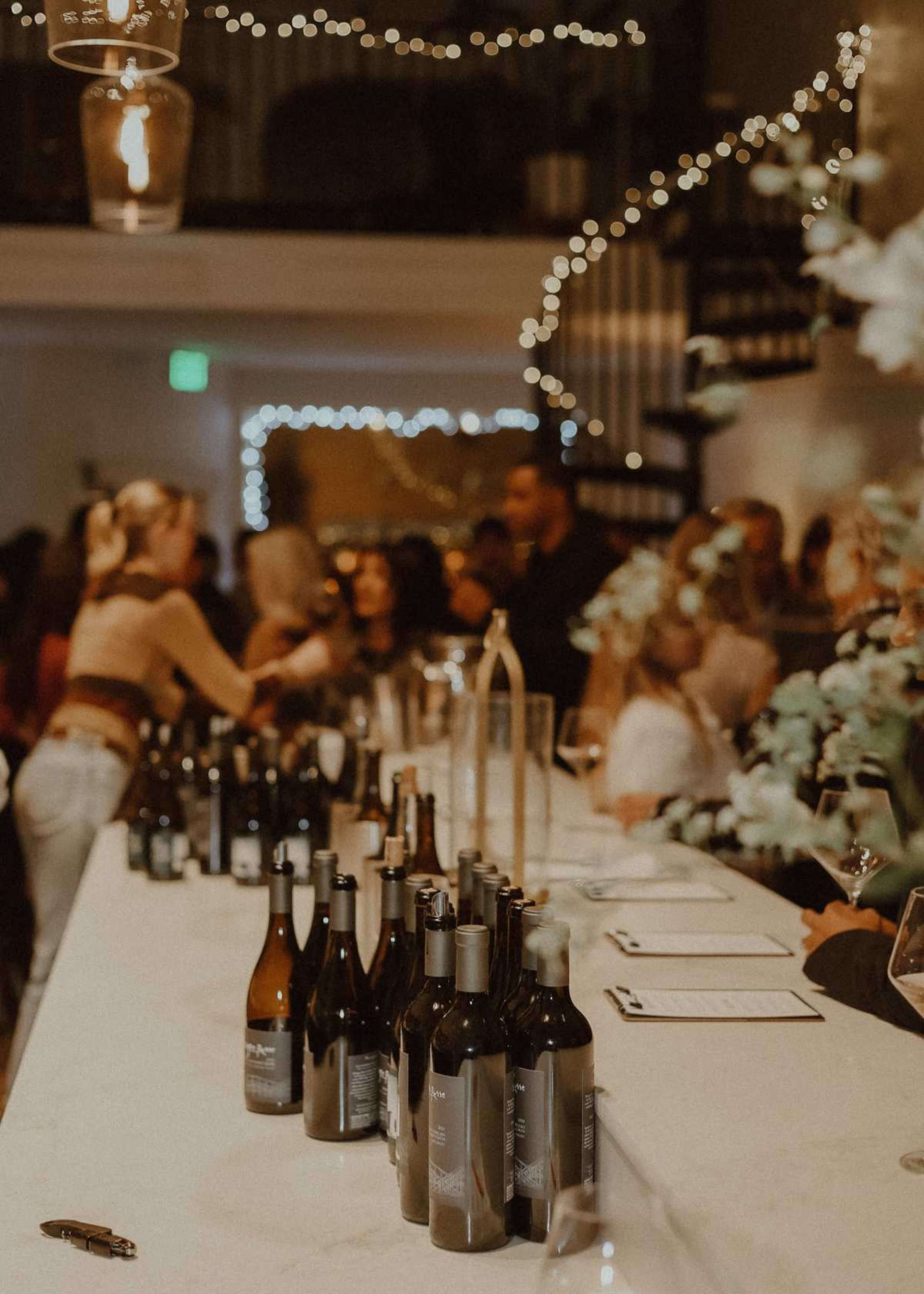 A bar area is set up with several bottles of wine lined up on a counter, while guests mingle in a dimly lit space adorned with string lights.