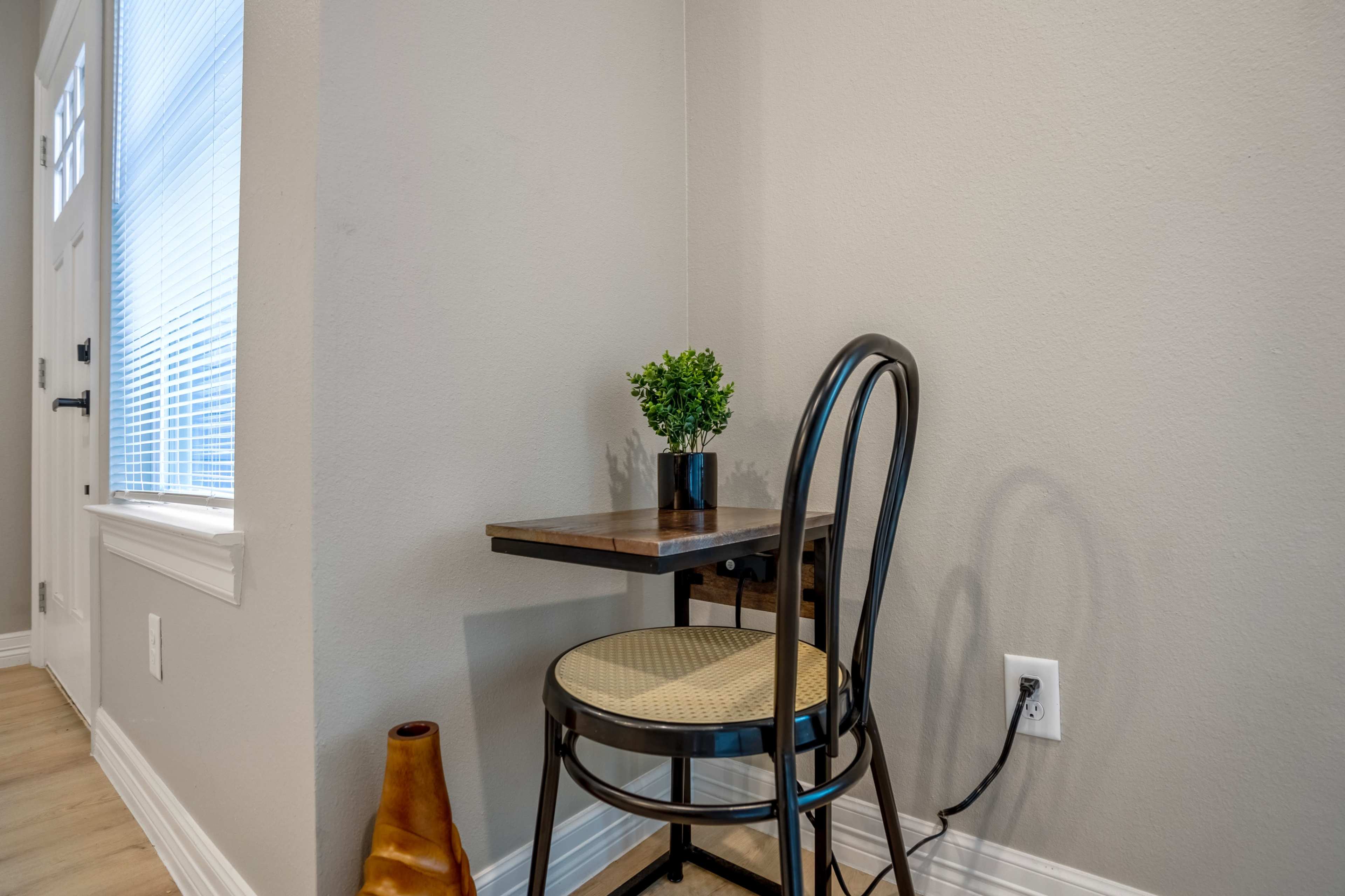 A small seating area features a wooden table in the corner with a chair and a potted plant on top, next to a wall with a power outlet.