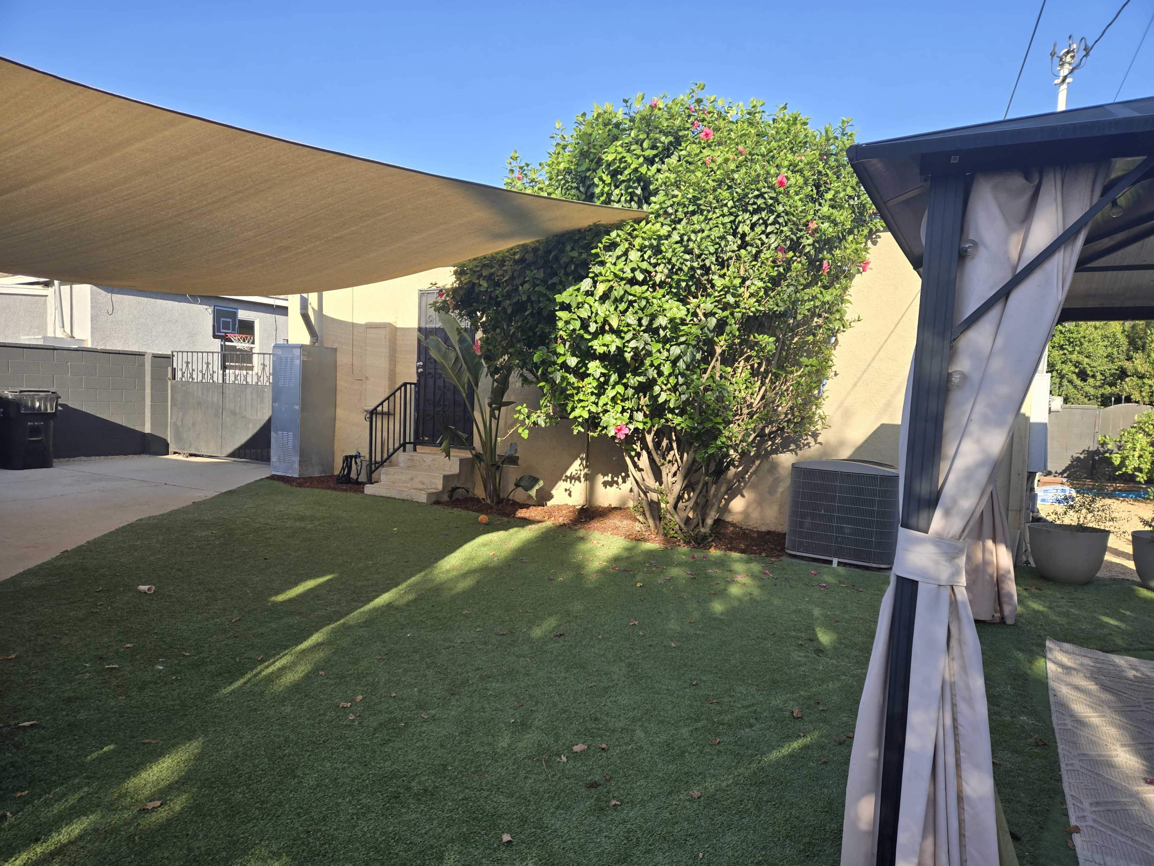 The image depicts a landscaped backyard with artificial grass, a shaded area provided by a fabric canopy, and a blooming shrub near a set of stairs.