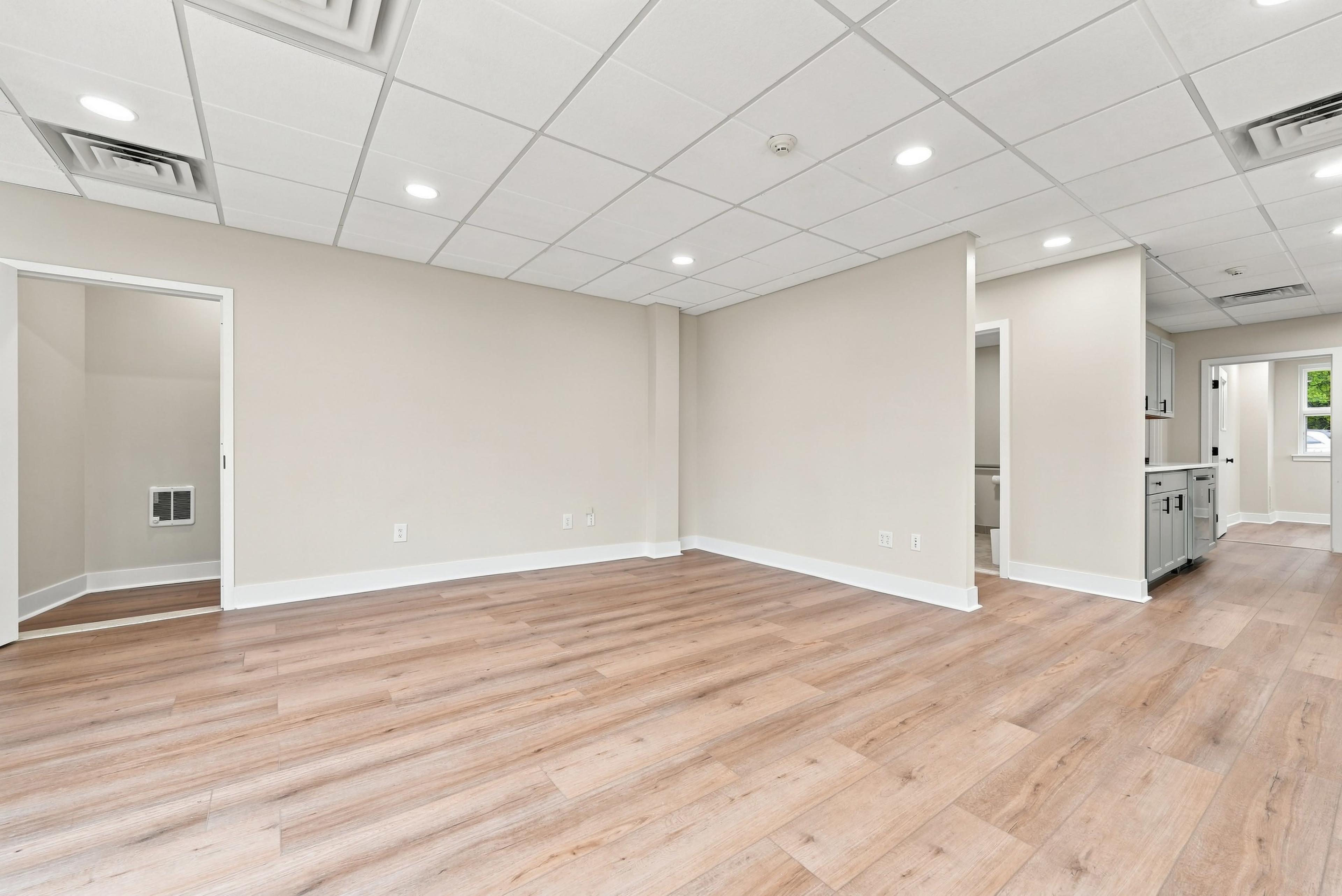 The image shows a spacious, unfurnished room with light-colored walls, a wooden floor, and a ceiling with recessed lighting.