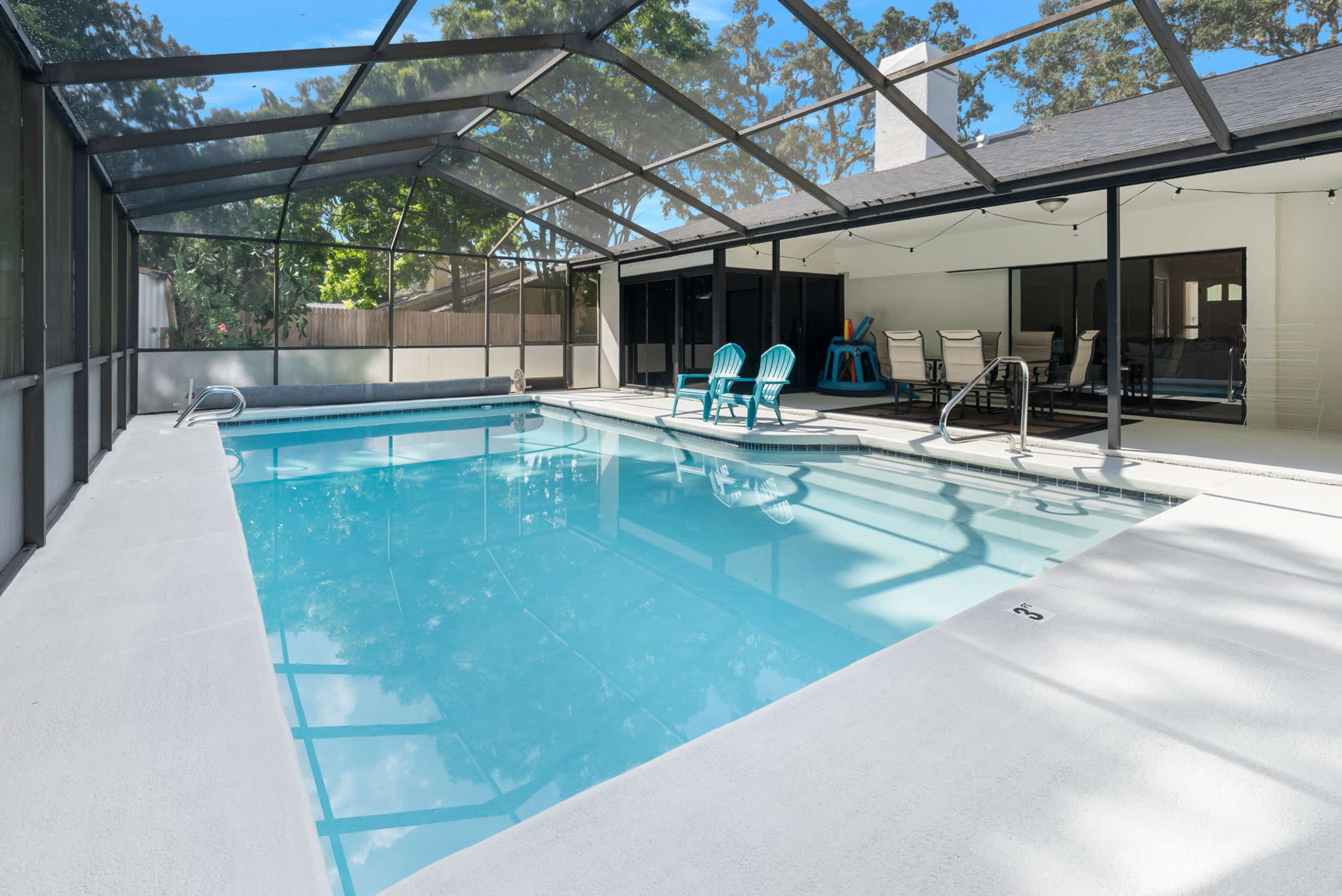 A clear swimming pool enclosed by a screened patio, flanked by lounge chairs and a covered seating area.