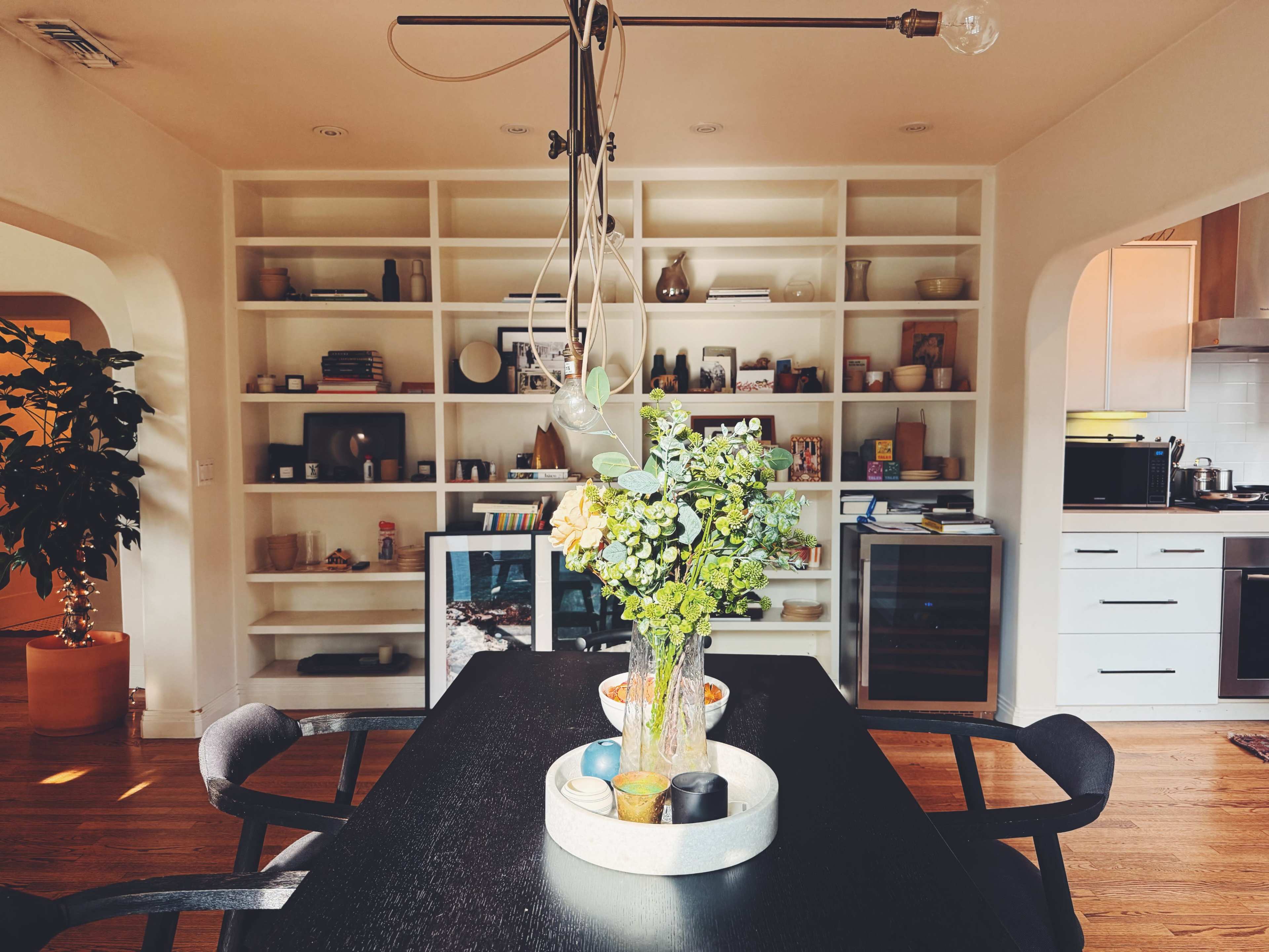 A dining area features a dark table with a vase of green flowers, surrounded by chairs, and a shelving unit filled with various items in the background.