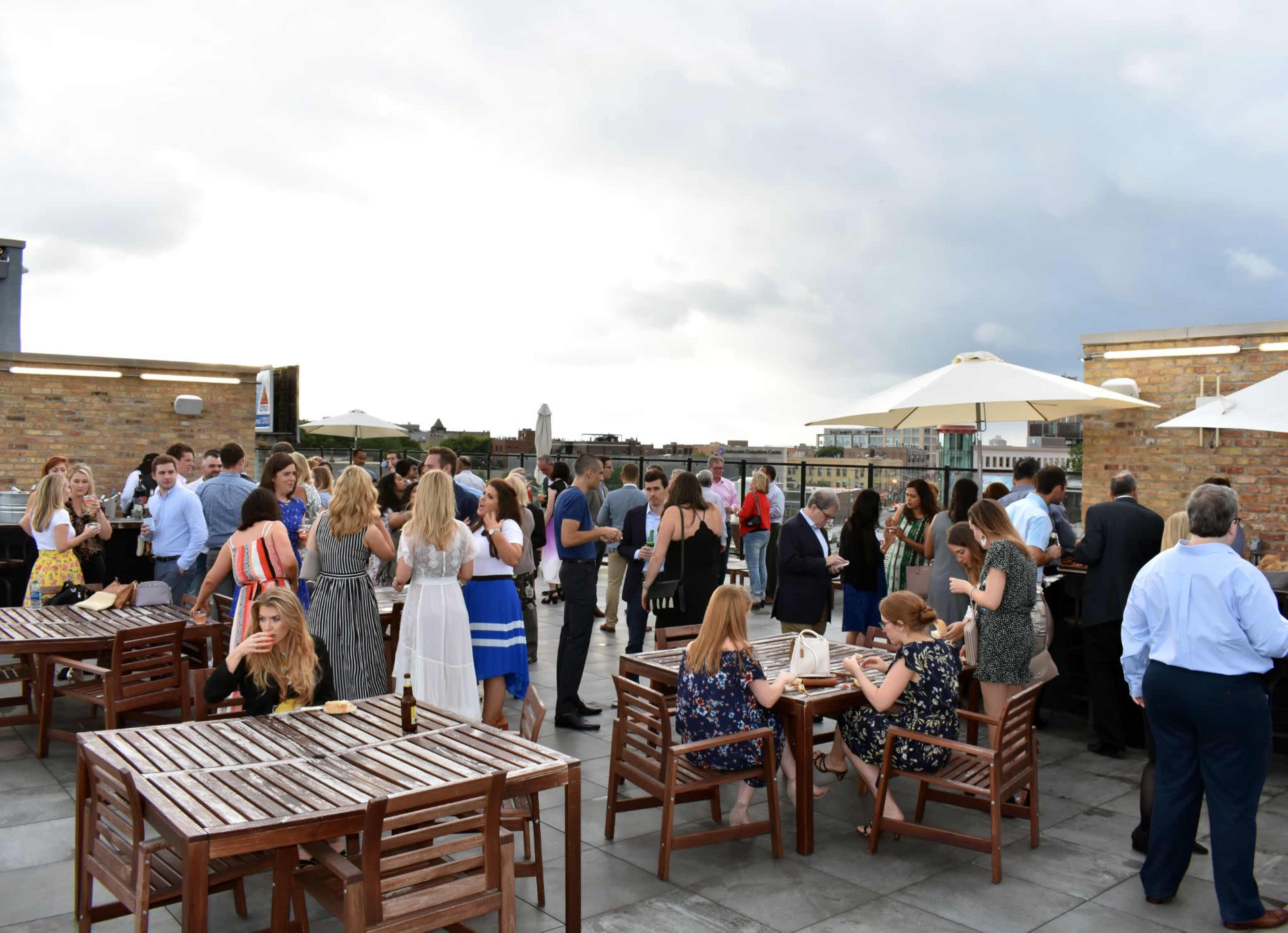 A large group of people mingles on a rooftop terrace under cloudy skies.