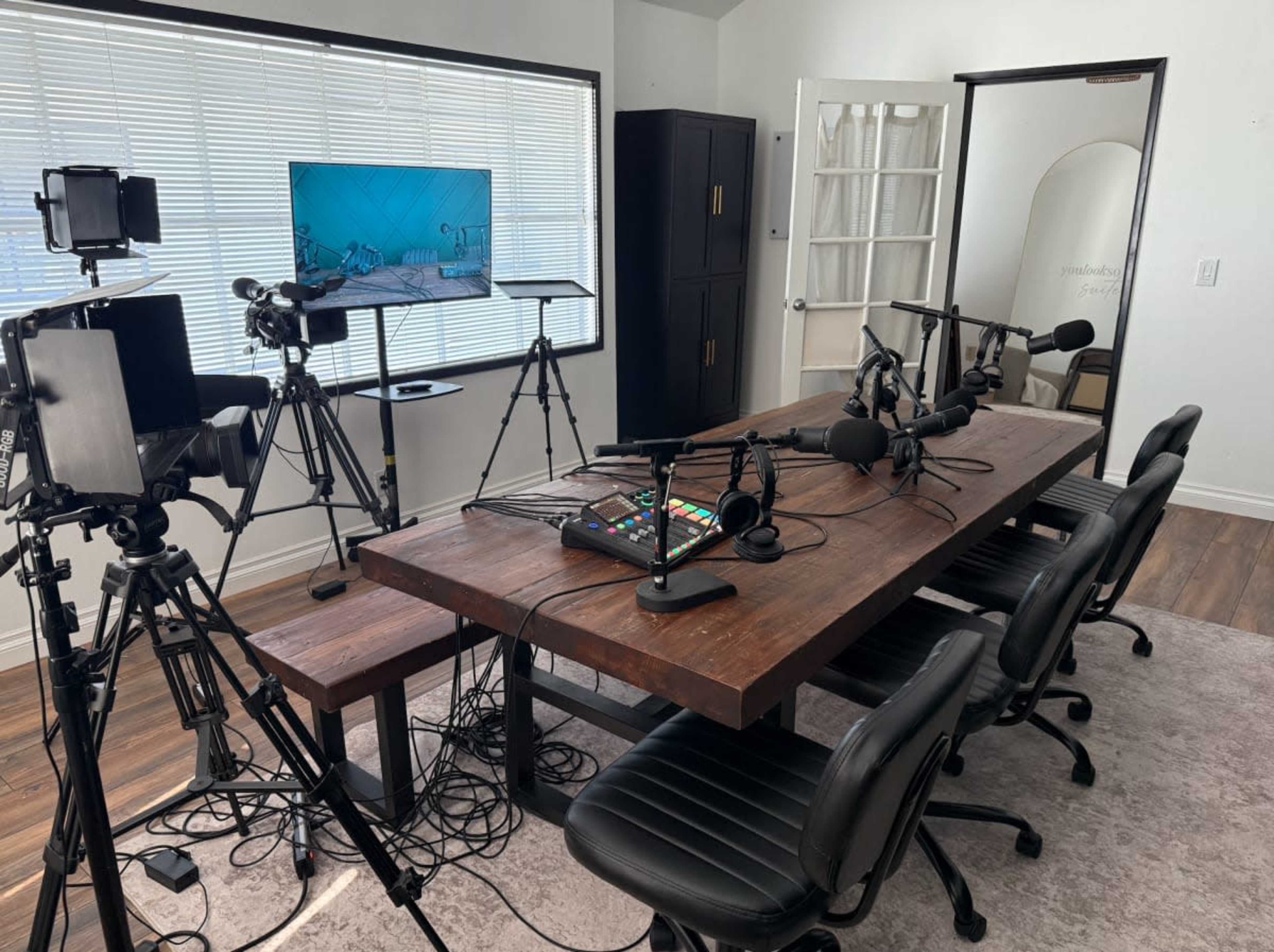 A video podcast studio is set up with a large wooden table, four black chairs, multiple microphones, cameras on tripods, and a monitor displaying a graphic in the background.