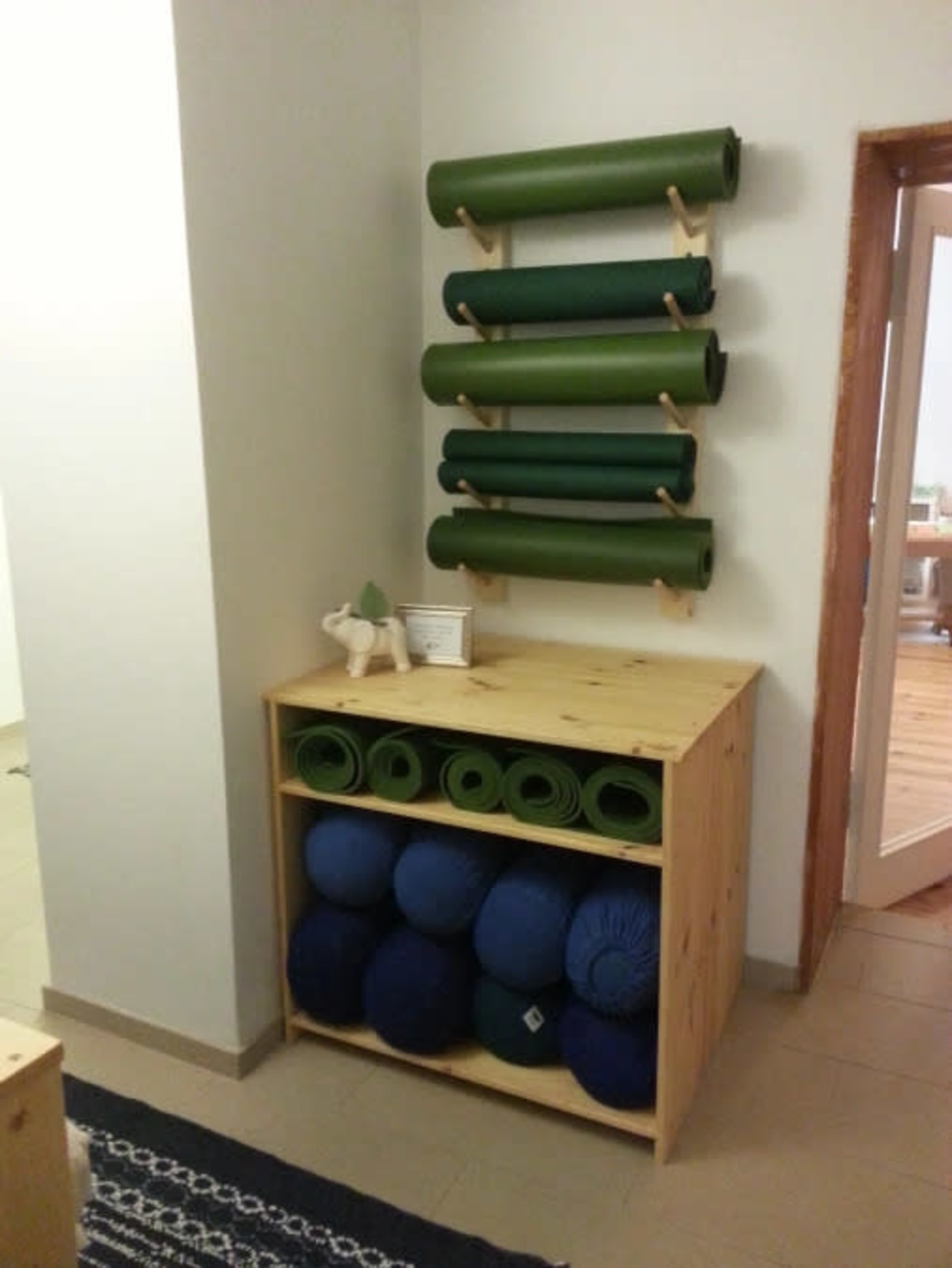 The image shows a wooden shelf mounted on a wall displaying rolled green yoga mats, with a storage unit below containing blue yoga bolsters and additional mats.