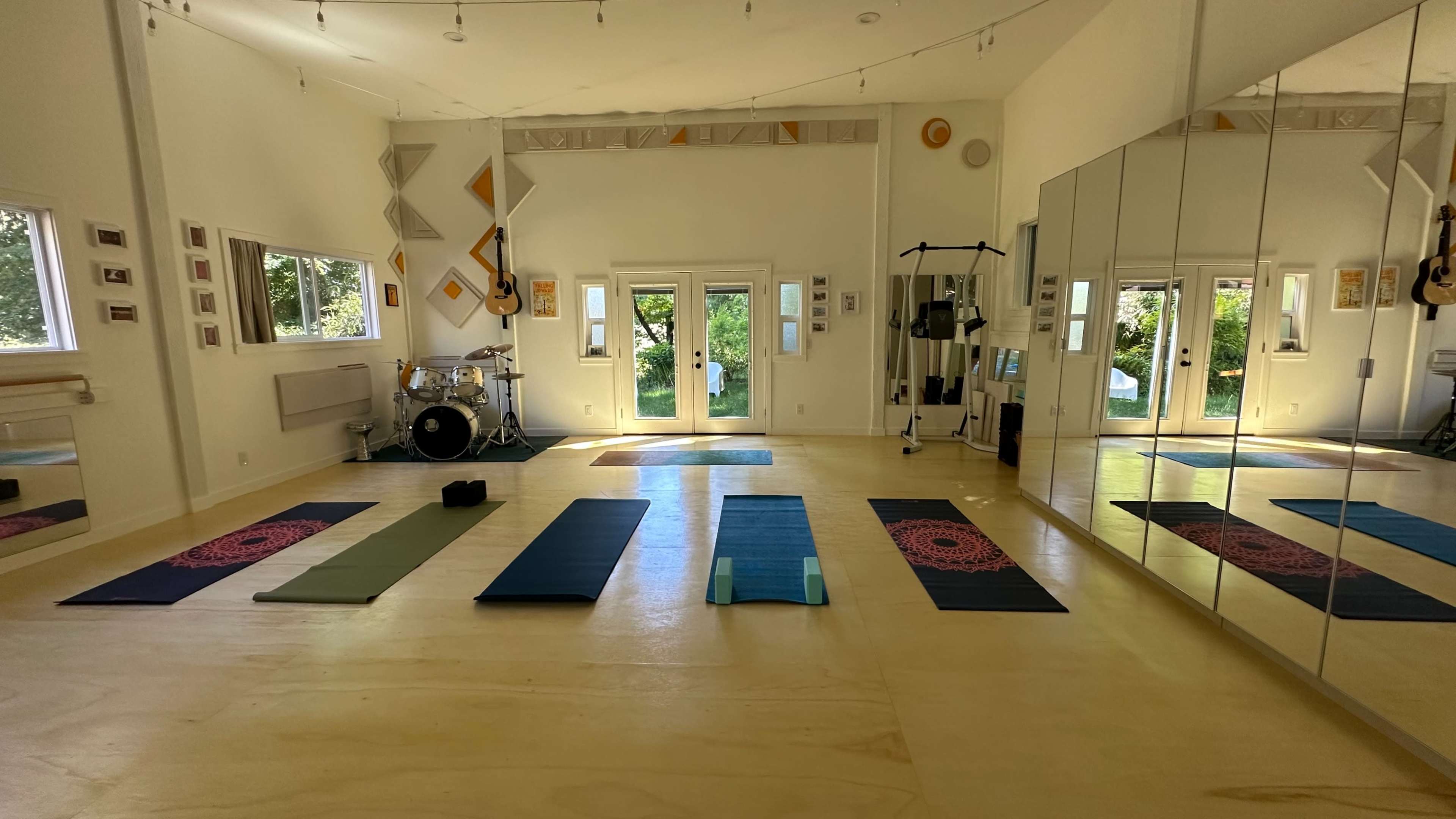 The image shows a spacious yoga studio with several mats laid out on a wooden floor, large windows allowing natural light, and a mirrored wall.