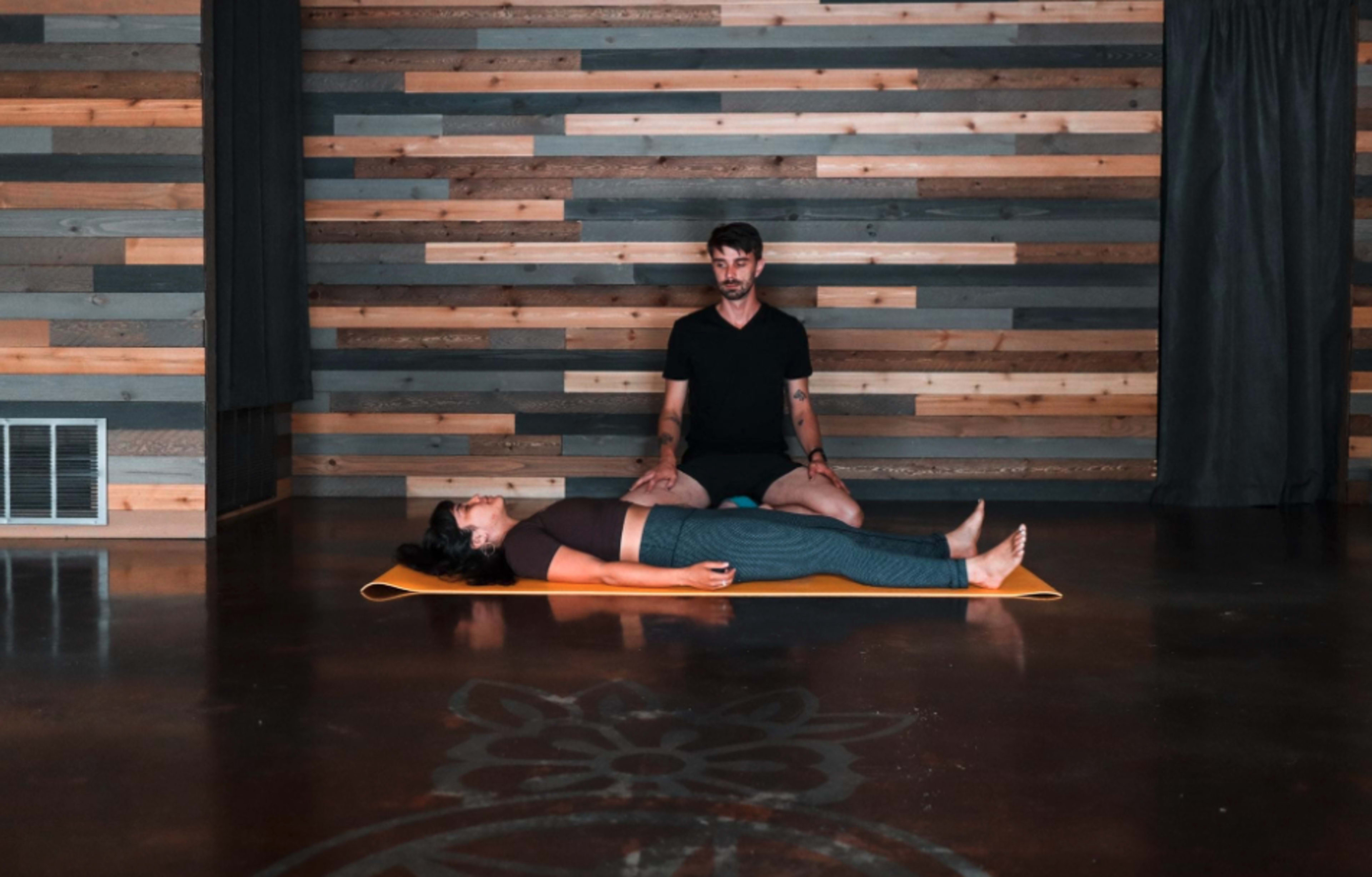 A person sits behind another person lying on a yoga mat in a studio with wooden walls.