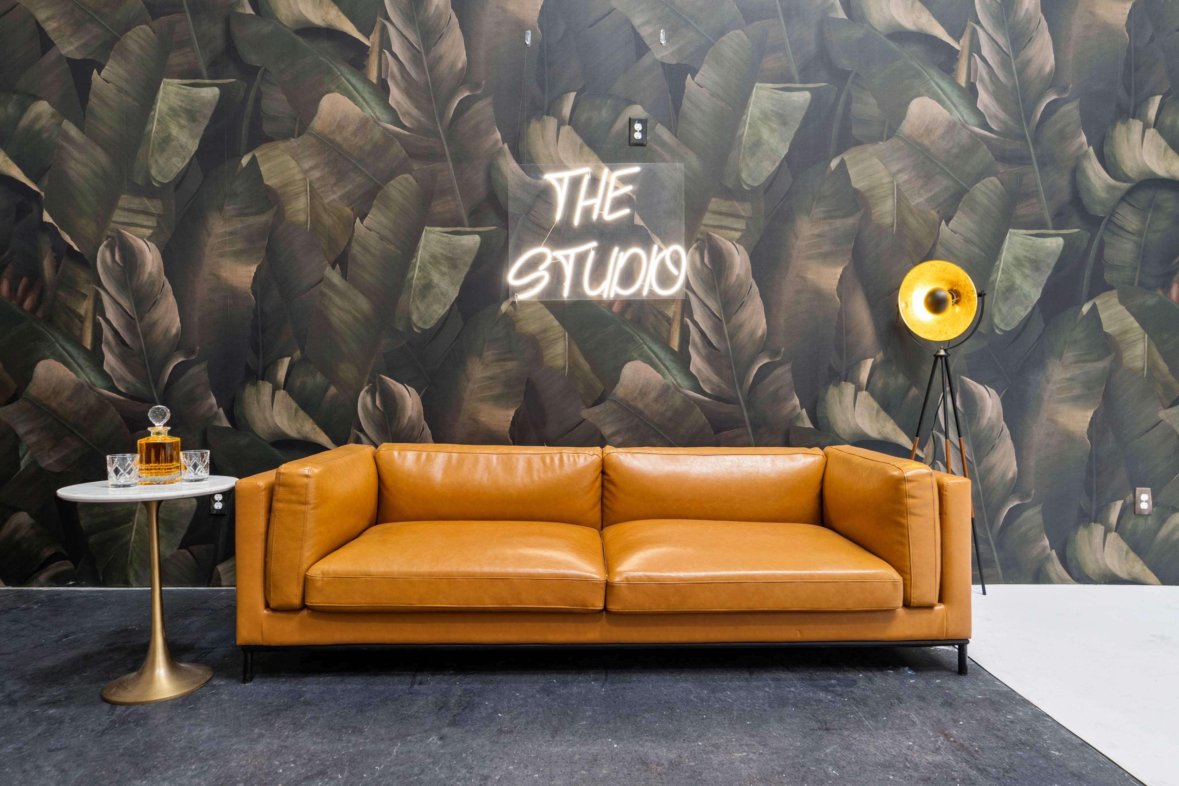A tan leather sofa is positioned in front of a leafy wallpaper, with a neon sign reading "THE STUDIO" and a vintage lamp beside a glass table with a decanter.