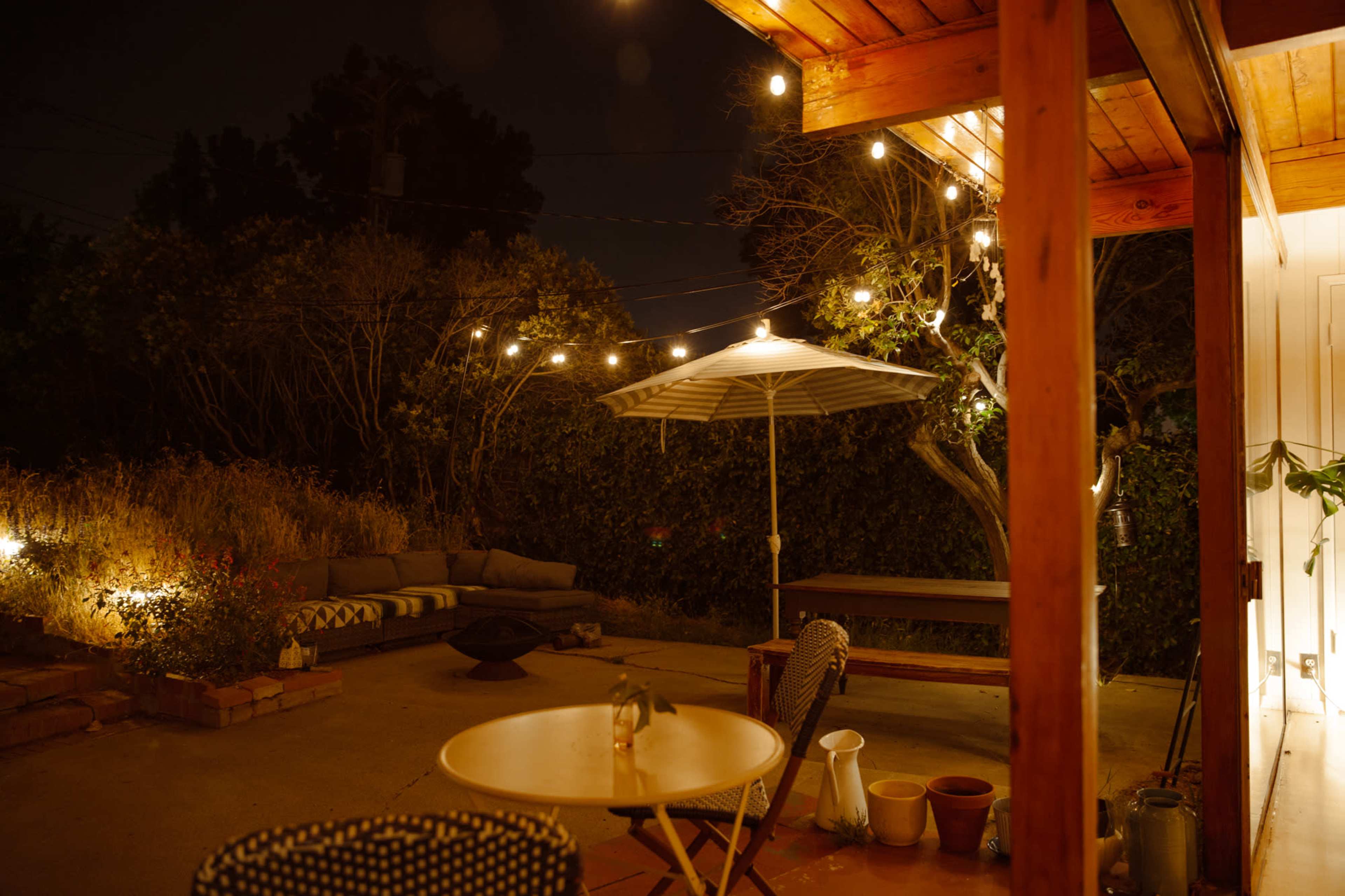 A cozy outdoor patio features string lights overhead, a shaded seating area, and various potted plants against a darkened backdrop.