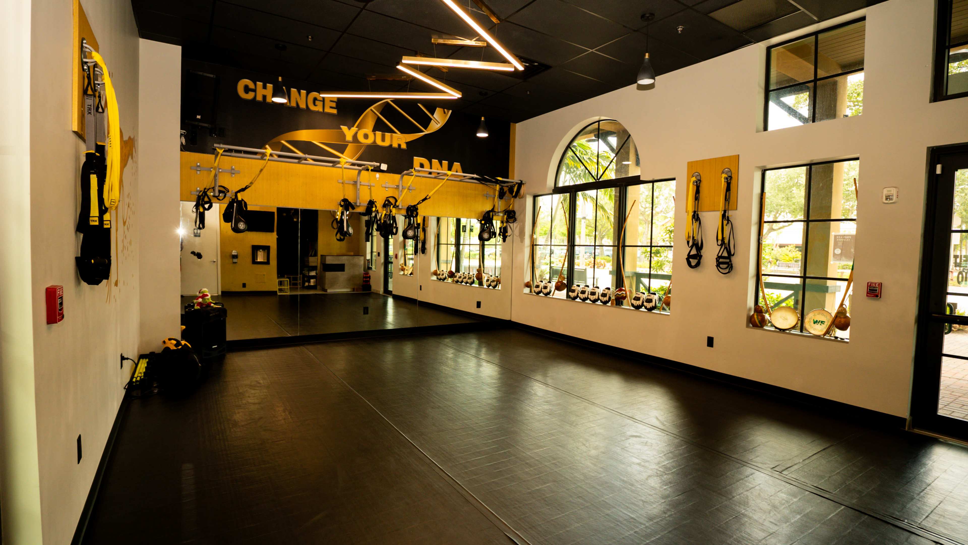 The image shows a well-lit fitness studio with mirrors, wall-mounted equipment, and a spacious black rubber floor.