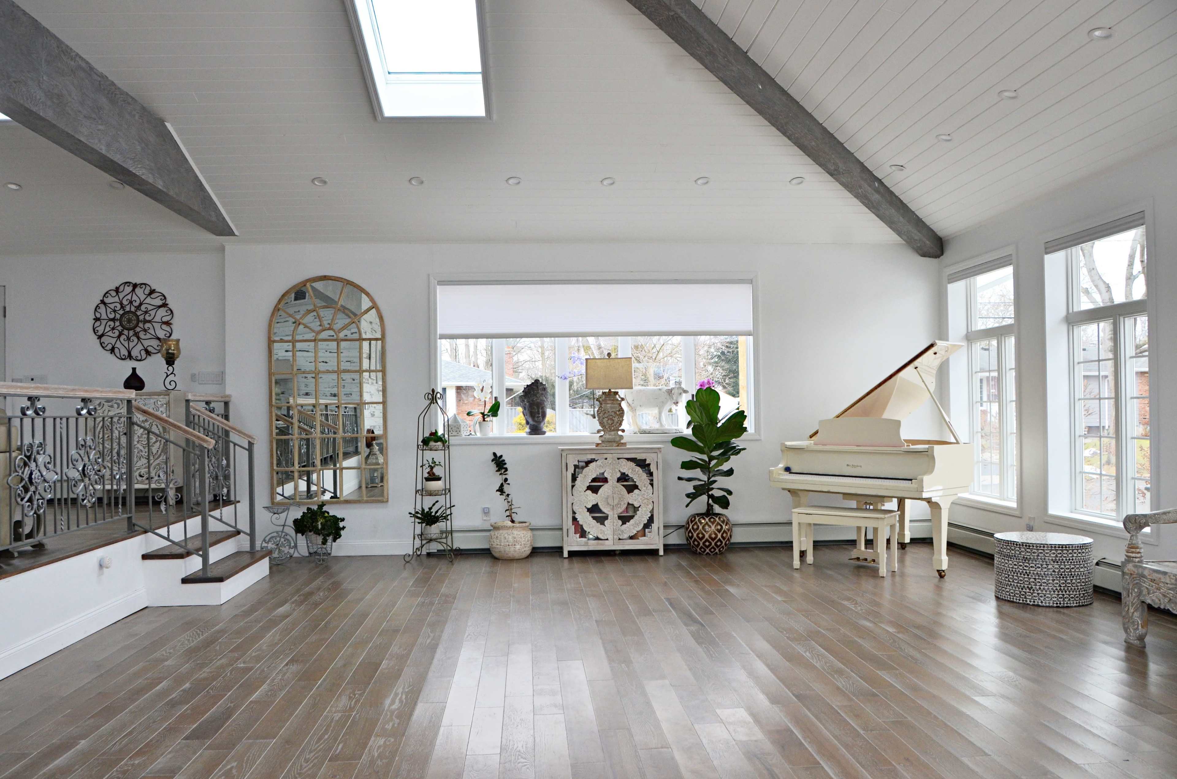 The spacious living room features hardwood flooring, large windows, a white piano, and decorative plants.