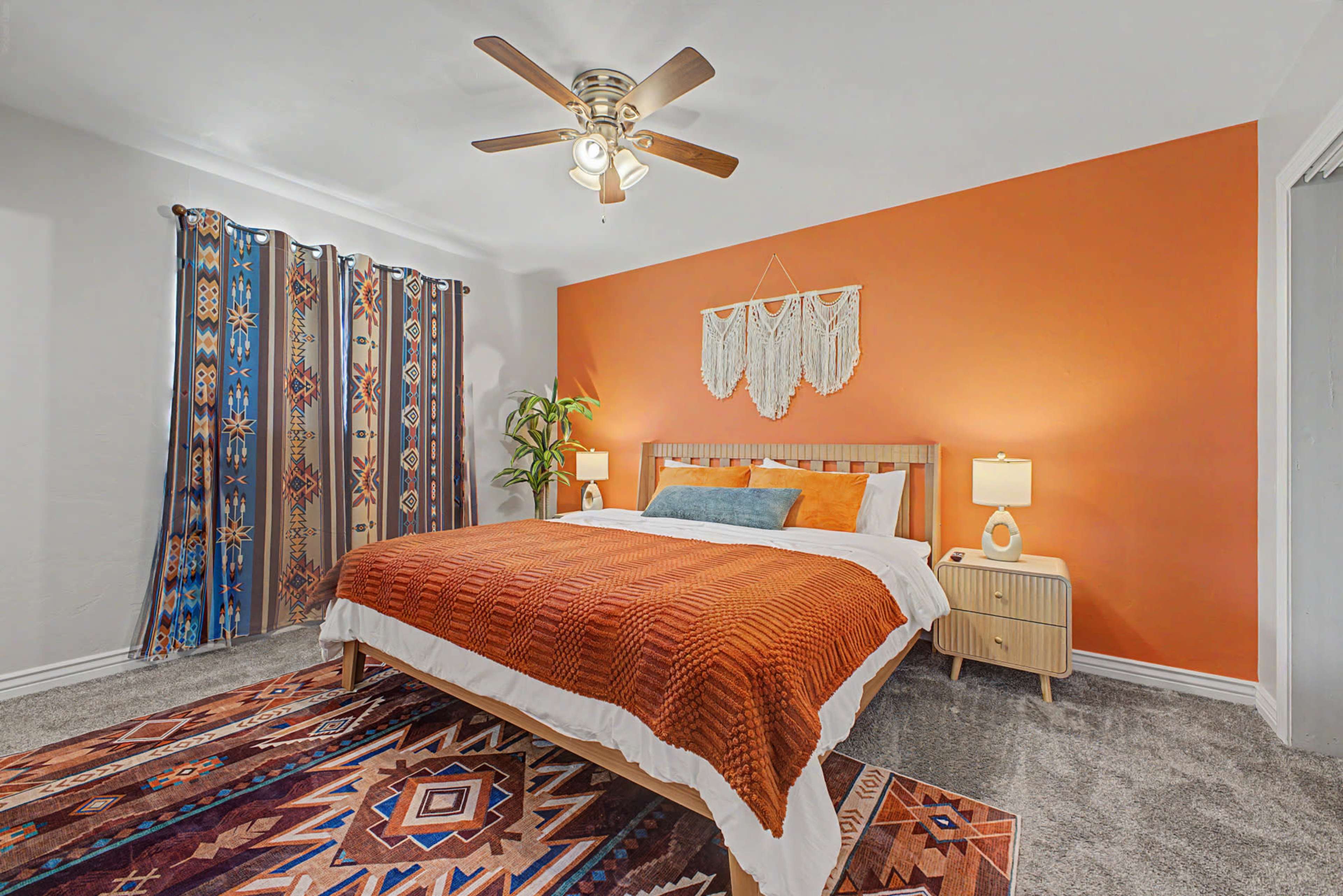 The image shows a cozy bedroom featuring an orange accent wall, a bed with an orange blanket, a patterned rug, and a ceiling fan.