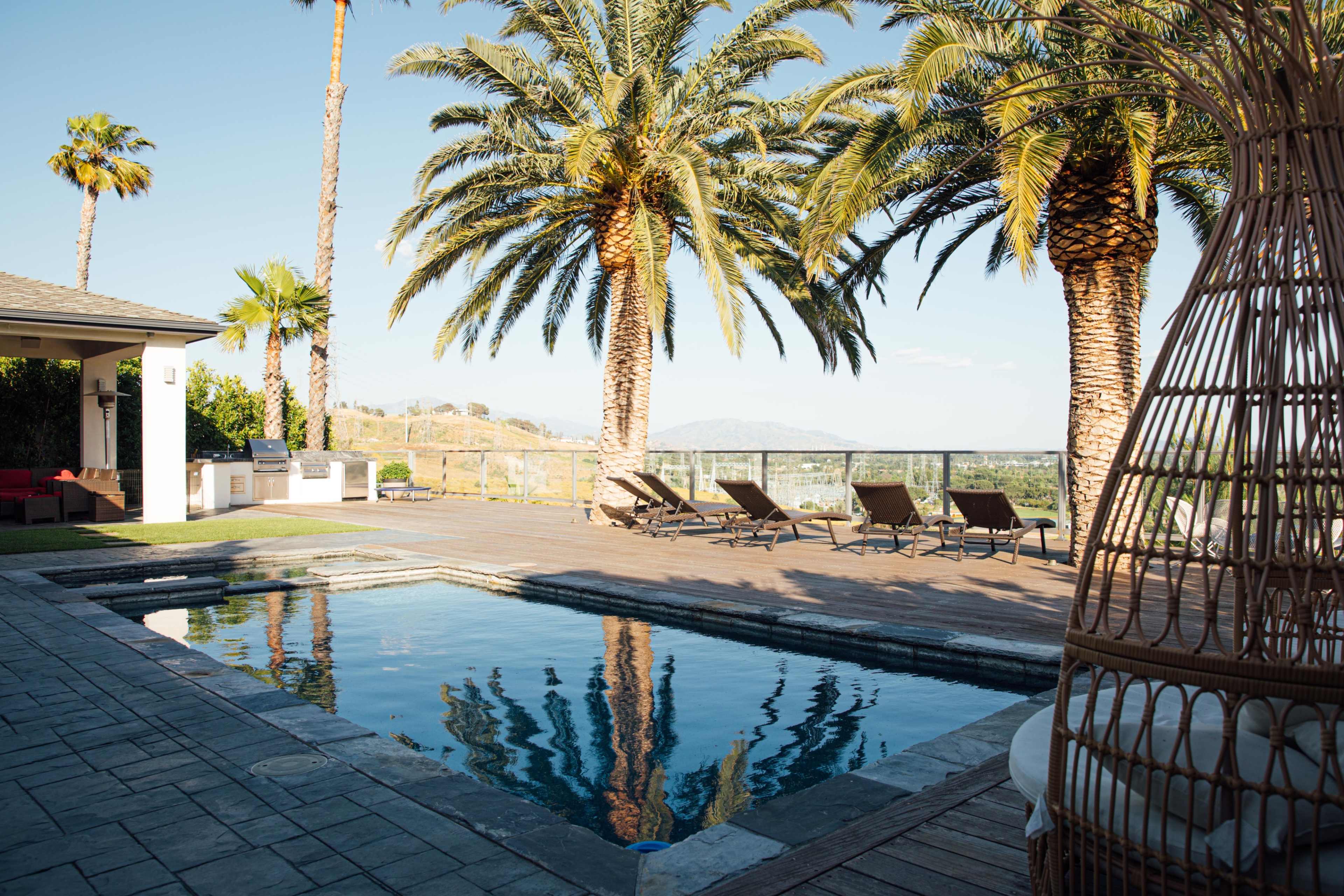 A poolside area surrounded by palm trees and lounge chairs, with a clear sky in the background.