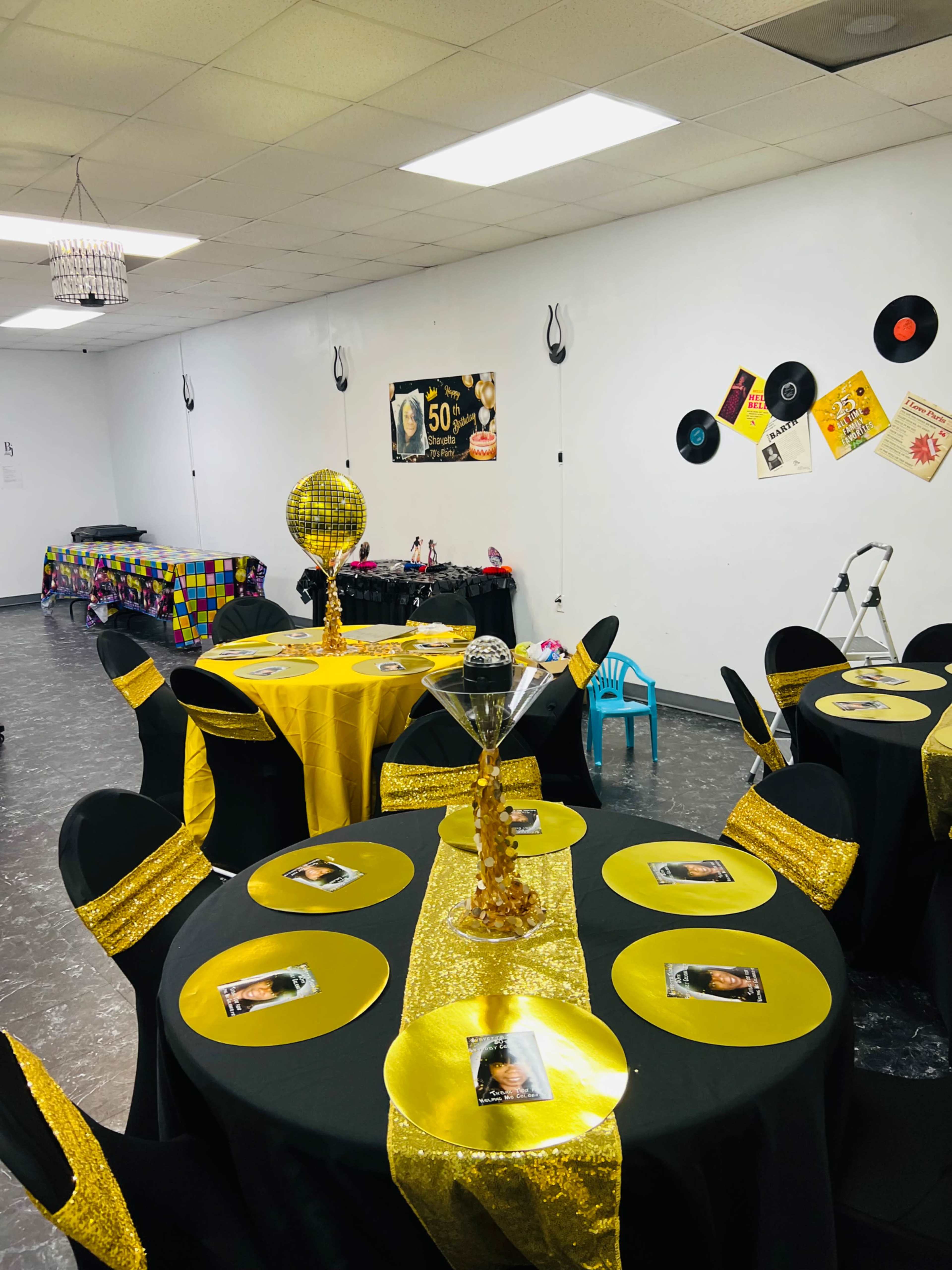 The image shows a party setup with round tables covered in black and gold tablecloths, featuring gold centerpieces and place settings, against a backdrop of party decor, including records and a banner.