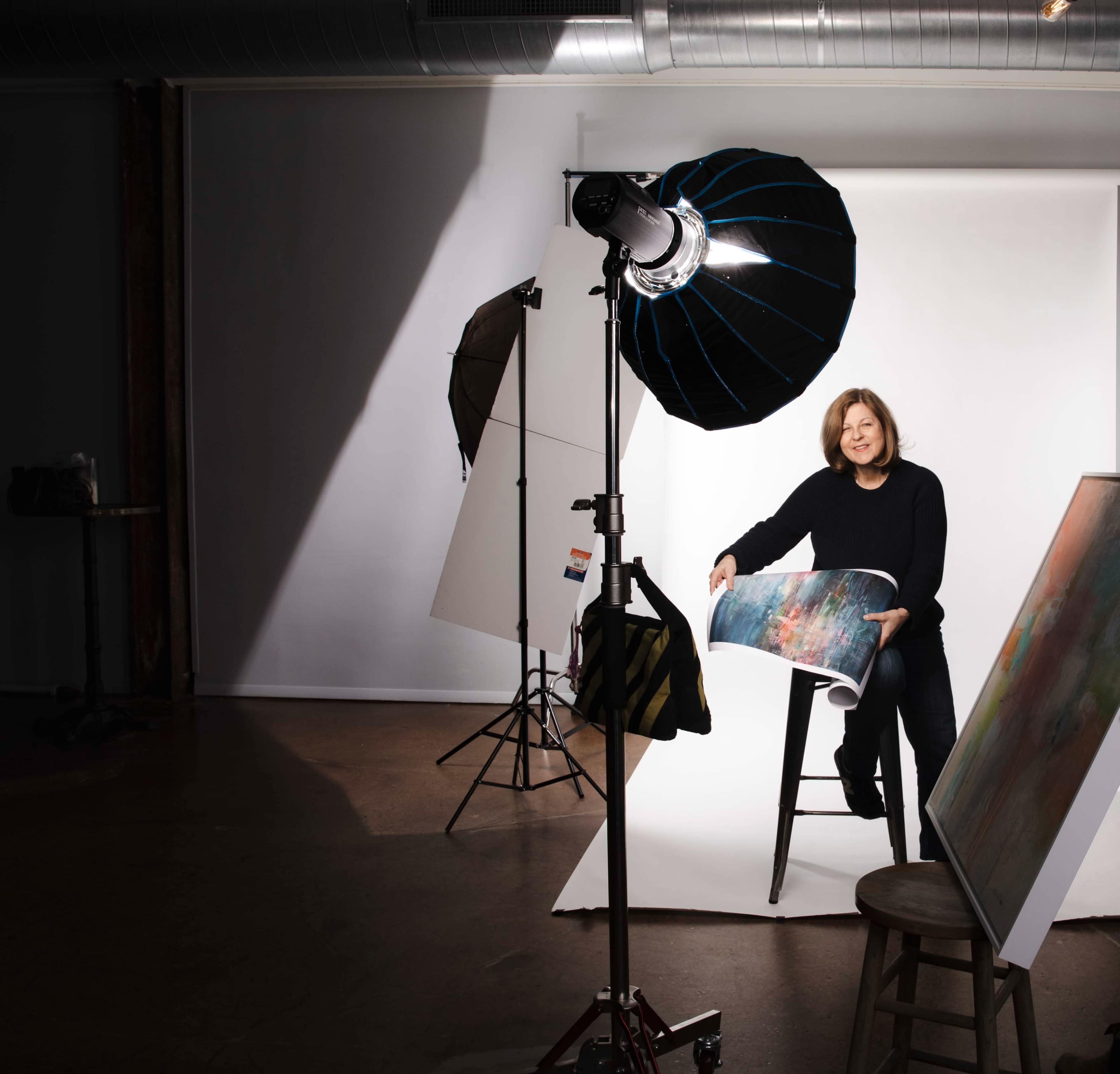 A woman stands beside a stool holding a rolled-up artwork in a well-lit photography studio.