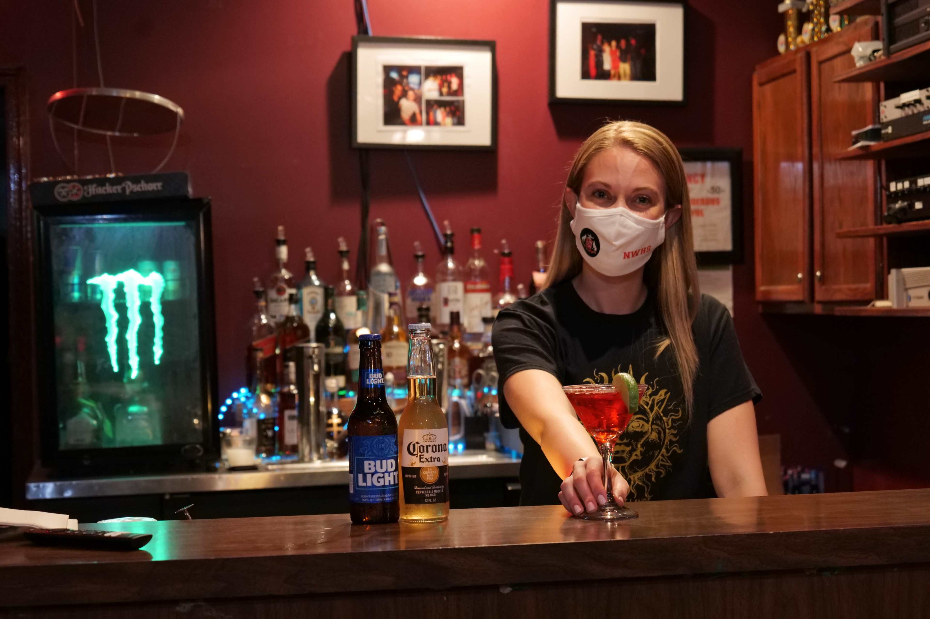 A bartender wearing a mask stands behind a bar, holding a cocktail while two beer bottles are placed on the counter.