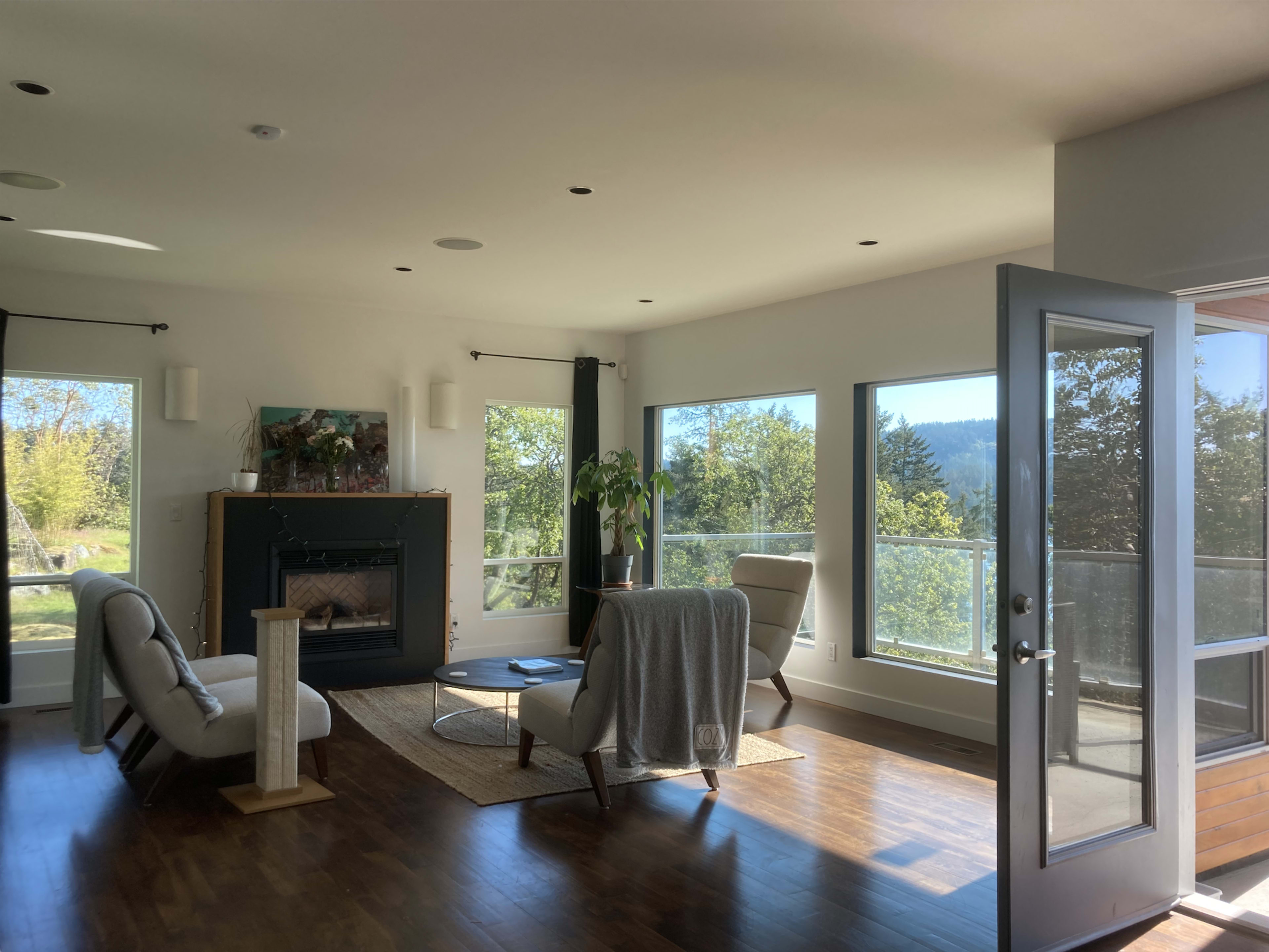 A modern living room features two chairs, a coffee table, and a fireplace, with large windows providing a view of the outdoors.