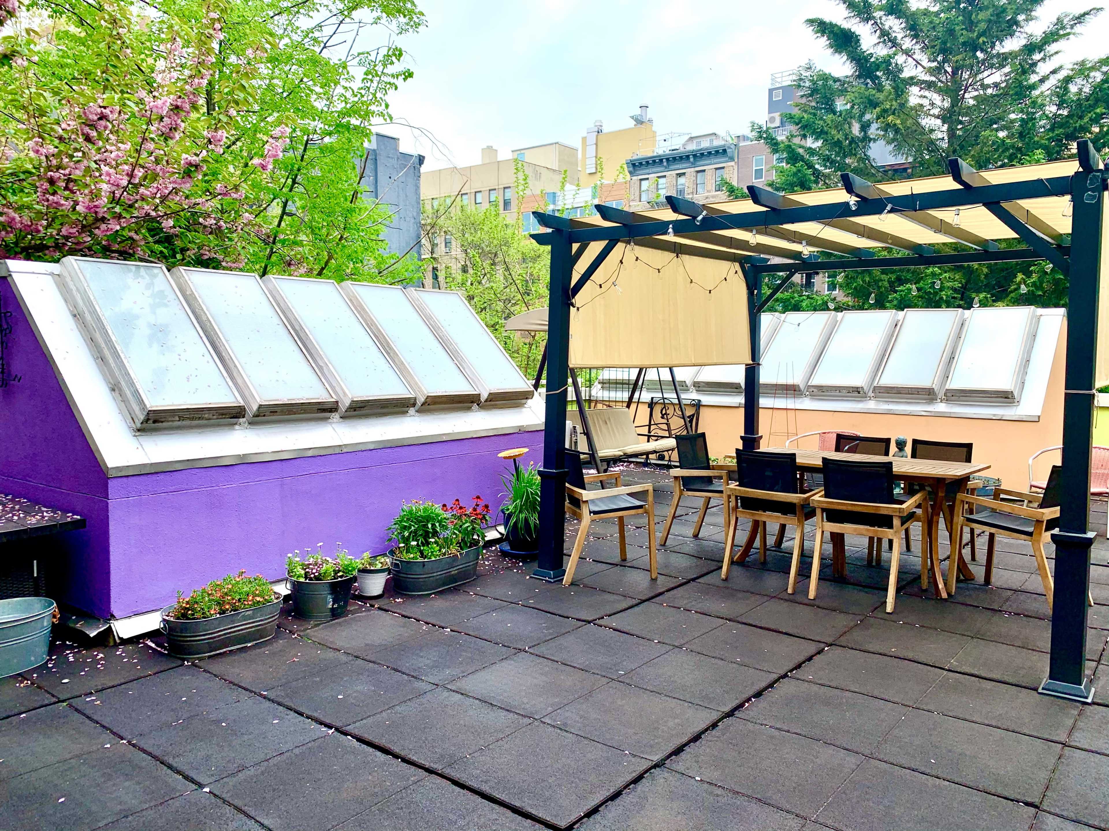 The image shows a rooftop space featuring purple walls with large windows, a wooden pergola, and a dining table surrounded by chairs.