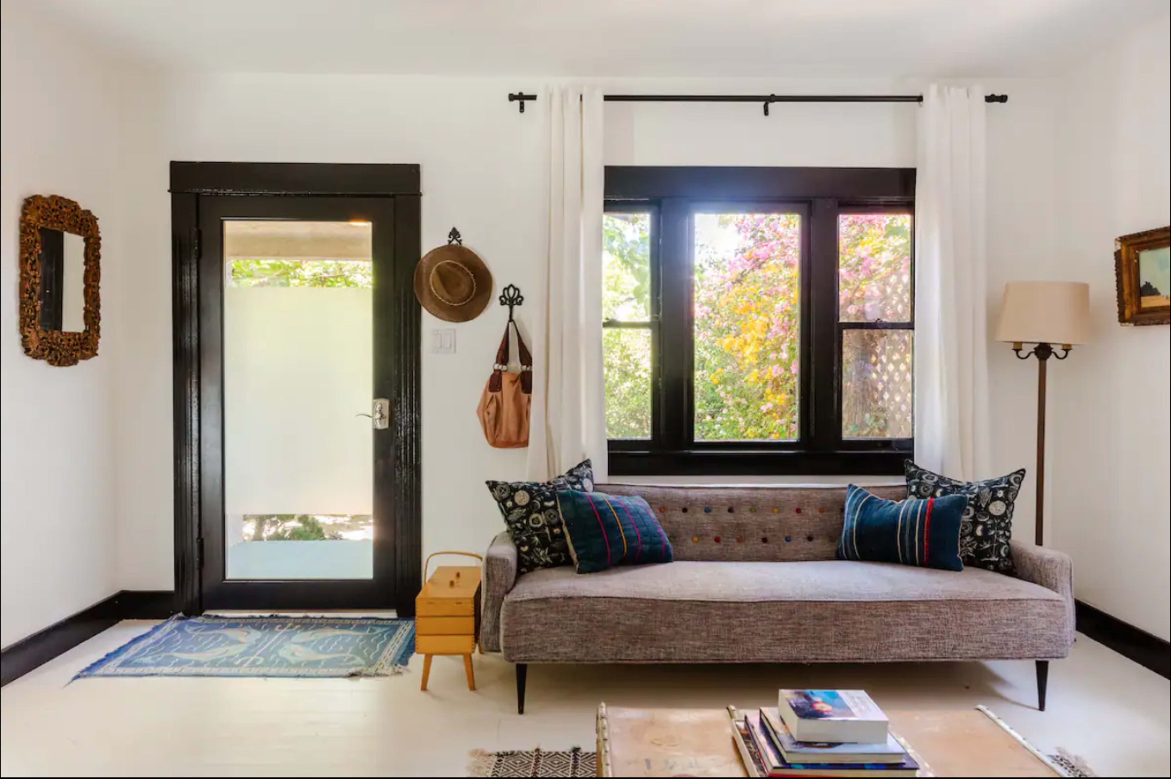 The image shows a cozy room featuring a brown couch with patterned cushions, a door leading outside, and large windows allowing natural light to enter.
