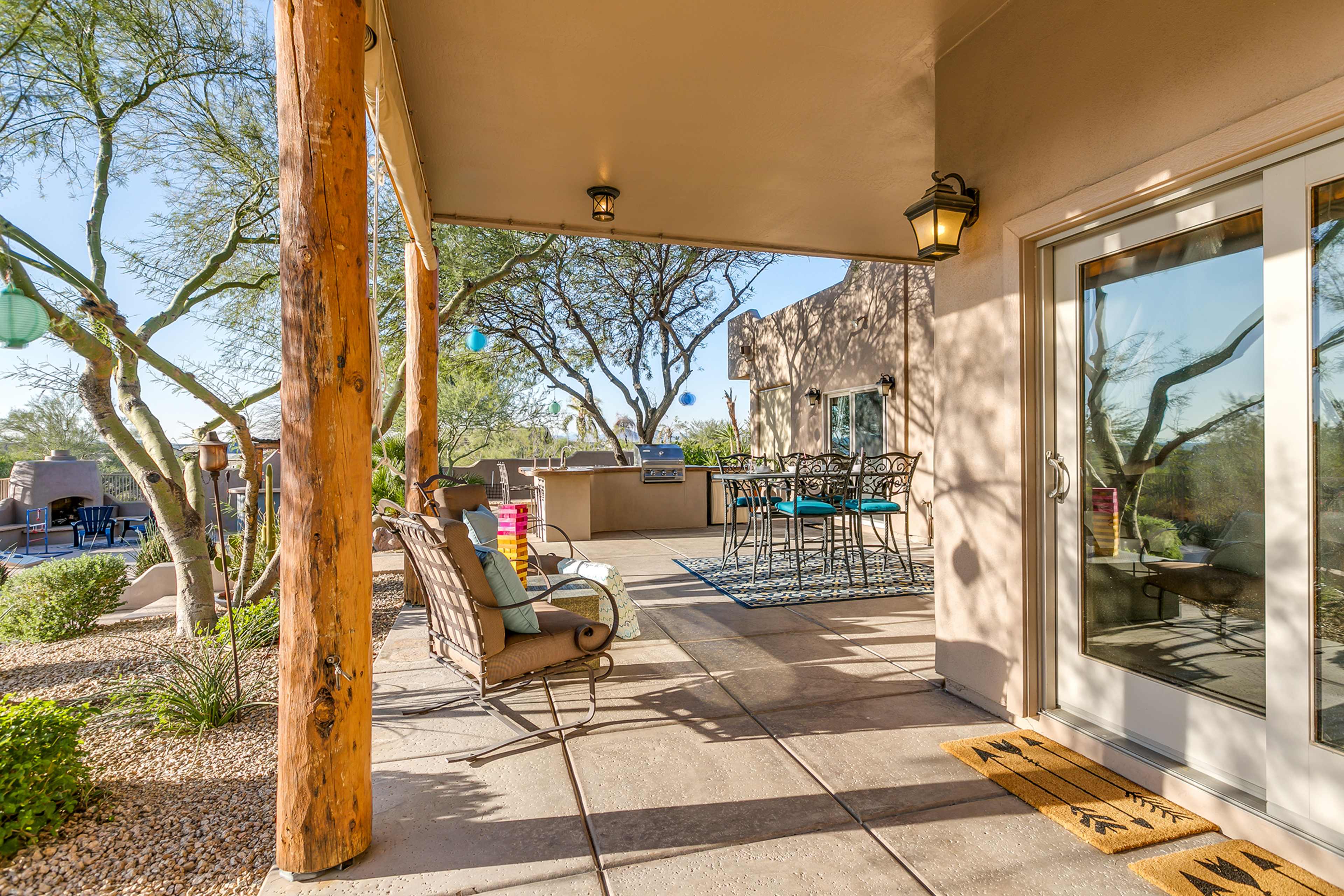 A patio area features a seating arrangement and dining set, surrounded by natural landscaping and structures in the background.