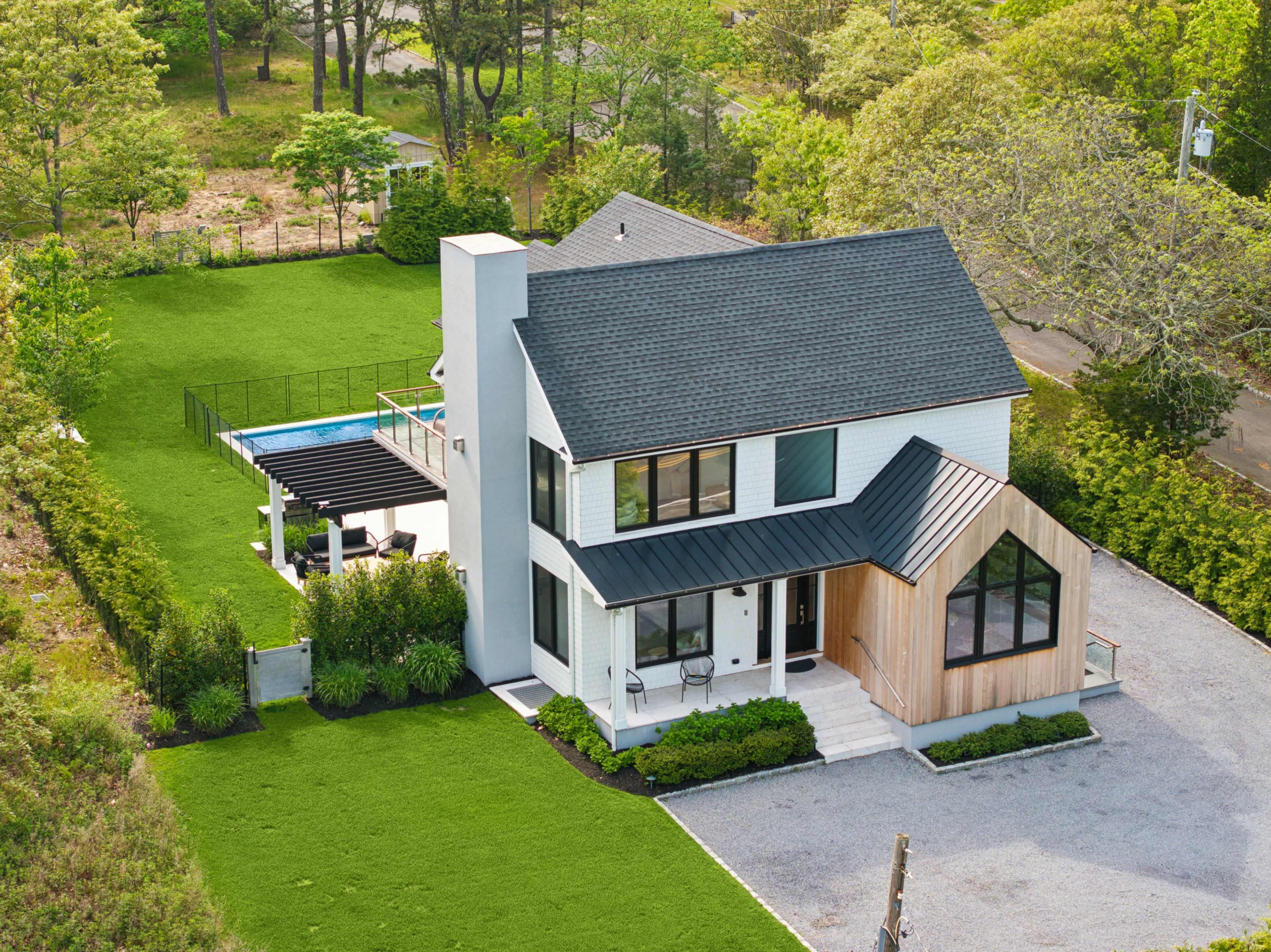 The image shows a modern two-story house with a blend of white siding and wooden accents, surrounded by well-maintained grass and landscaping, with a swimming pool visible in the backyard.