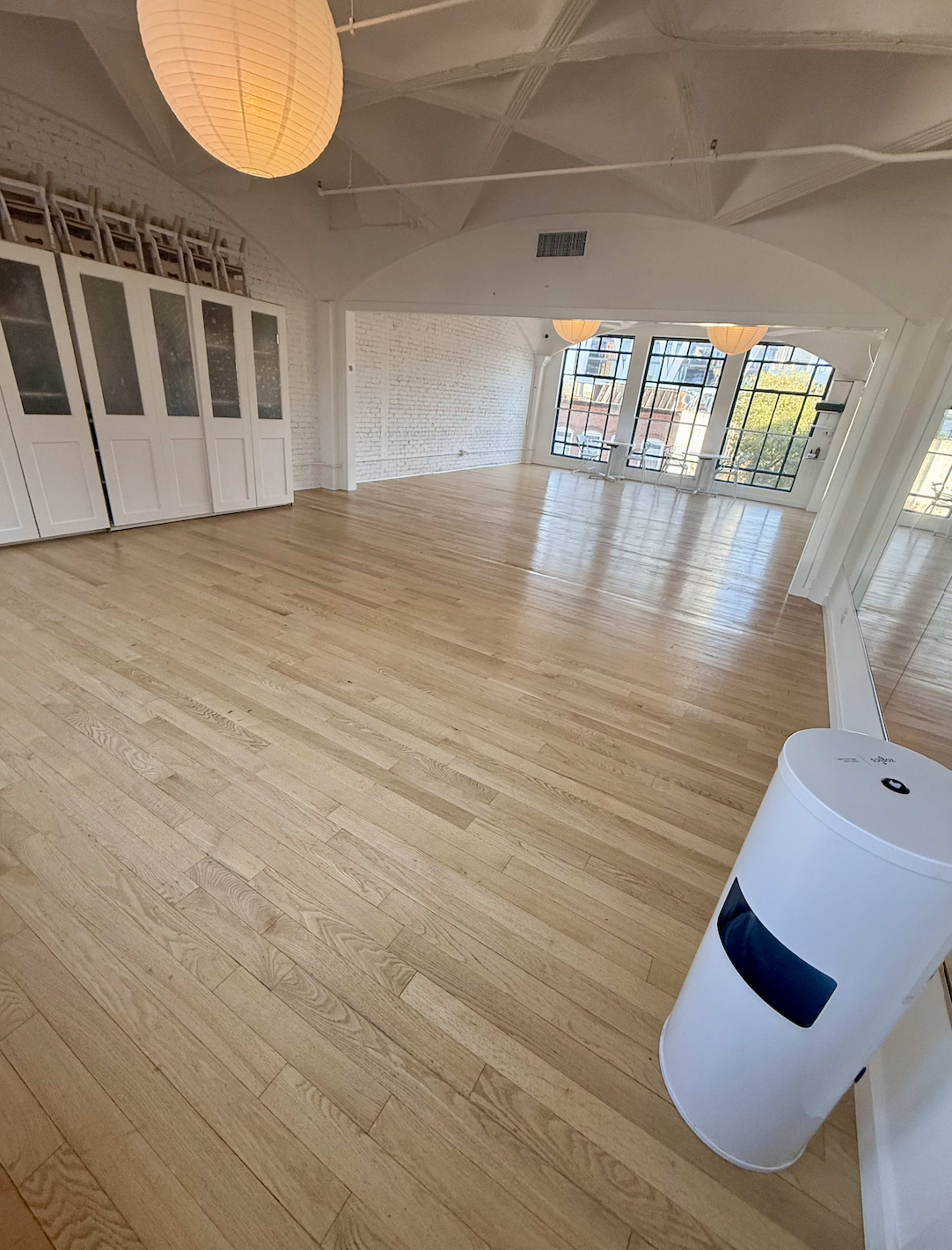 The image shows a spacious, well-lit room with hardwood floors, large windows, and a white storage cabinet along one wall.