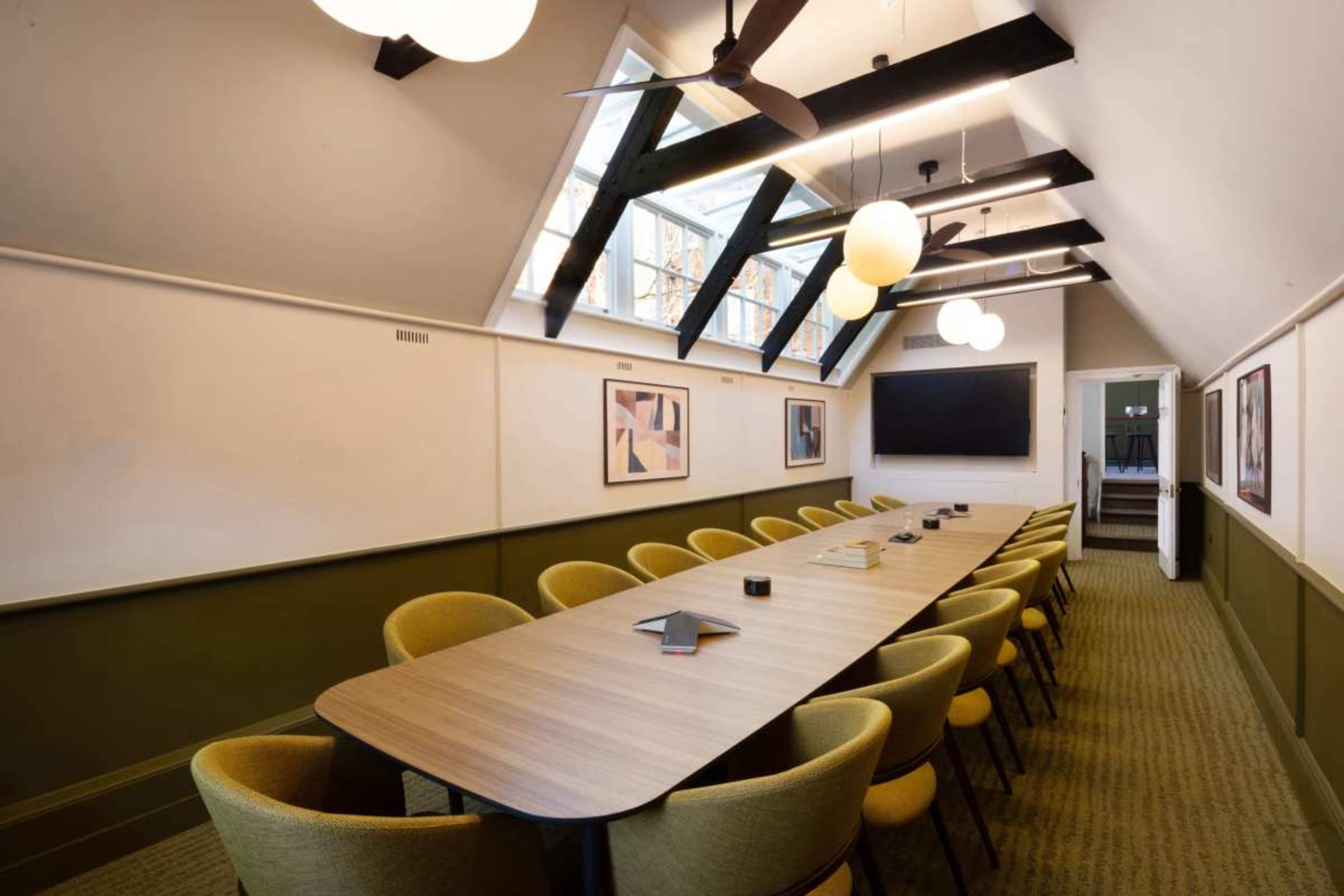 The image shows a modern conference room with a long wooden table surrounded by yellow chairs, featuring large windows and unique ceiling beams.