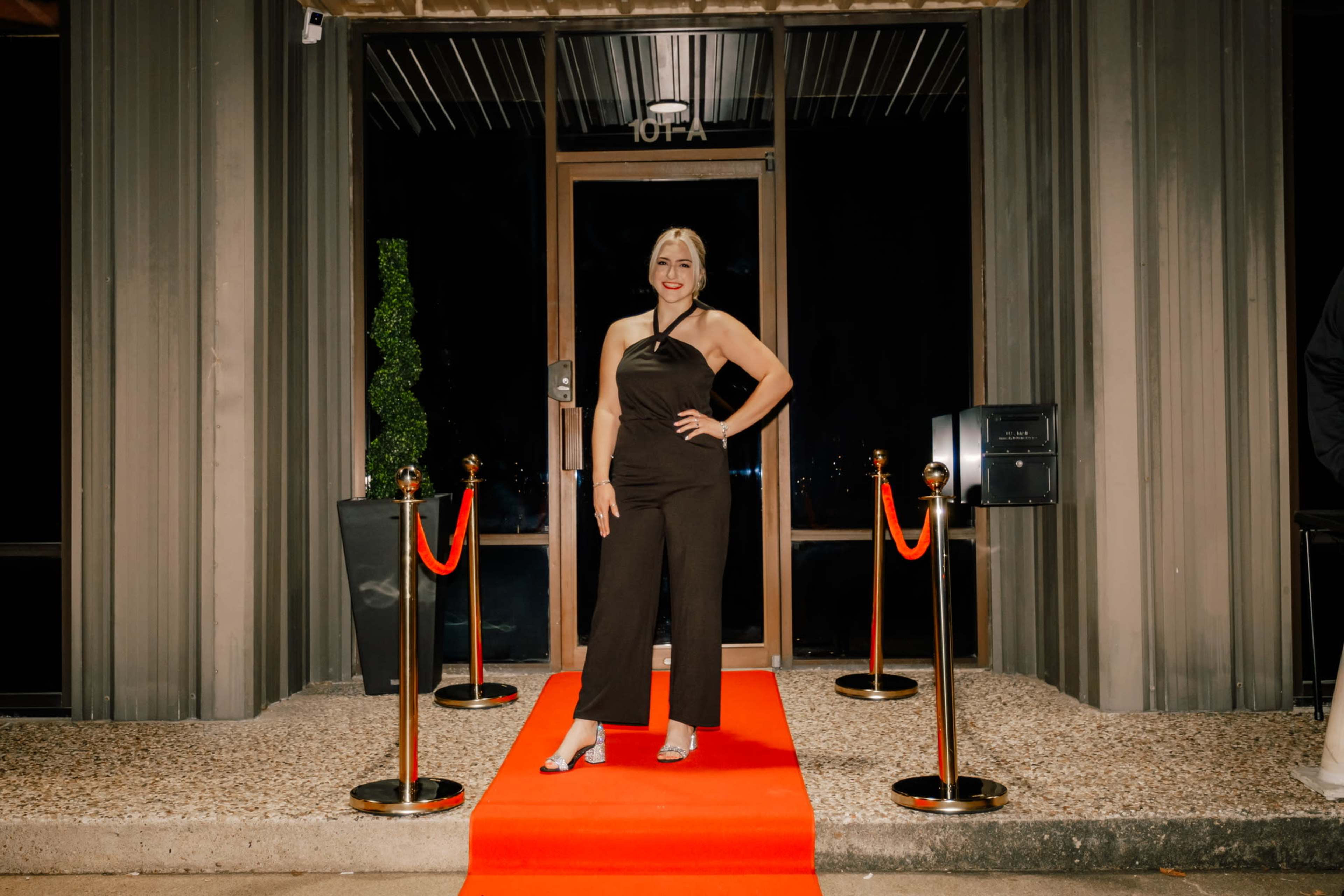 A woman stands on a red carpet in front of a building entrance adorned with golden stanchions and a mailbox.
