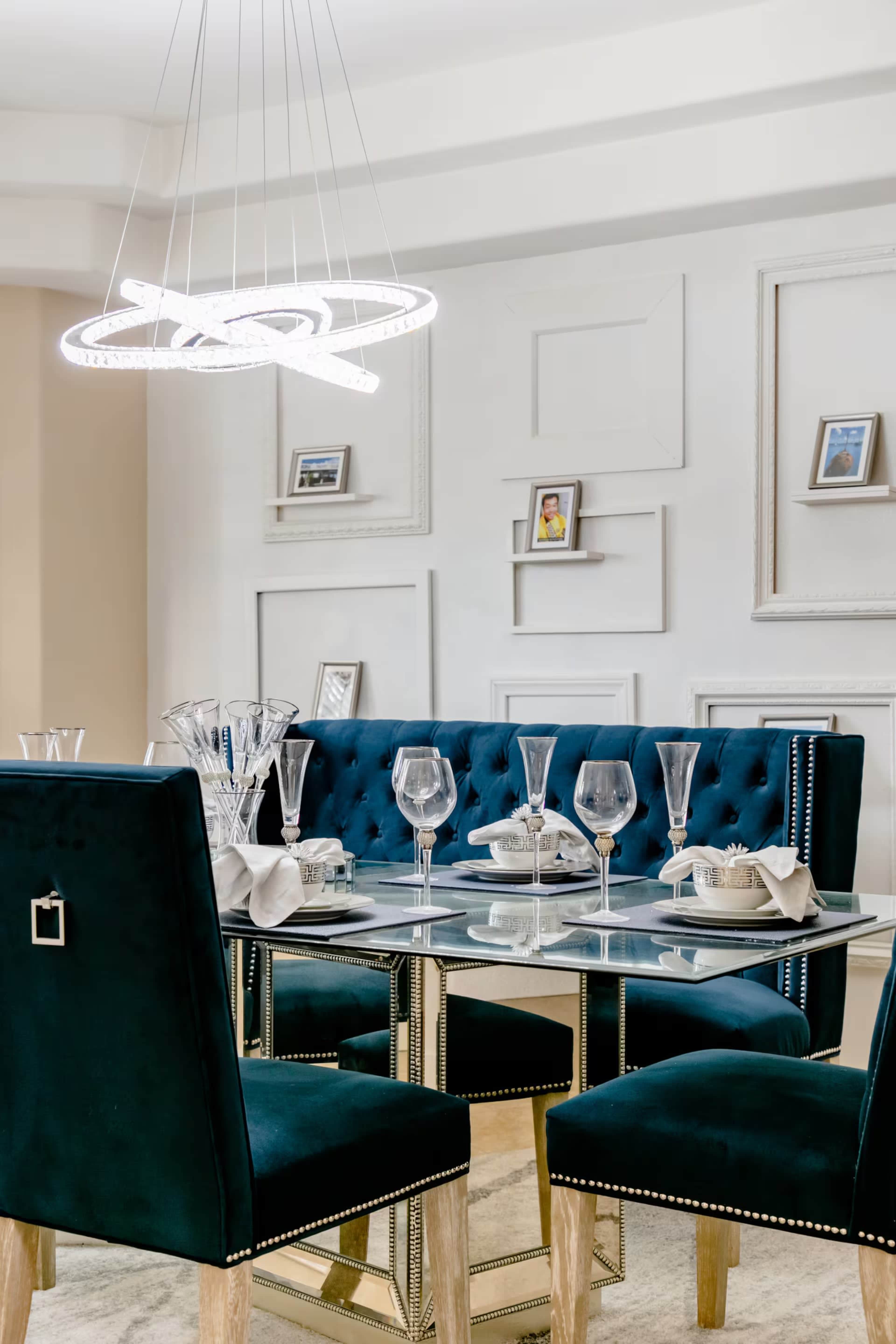 A modern dining room features a glass table with elegant tableware set for a meal, surrounded by stylish blue upholstered chairs and a contemporary light fixture overhead.